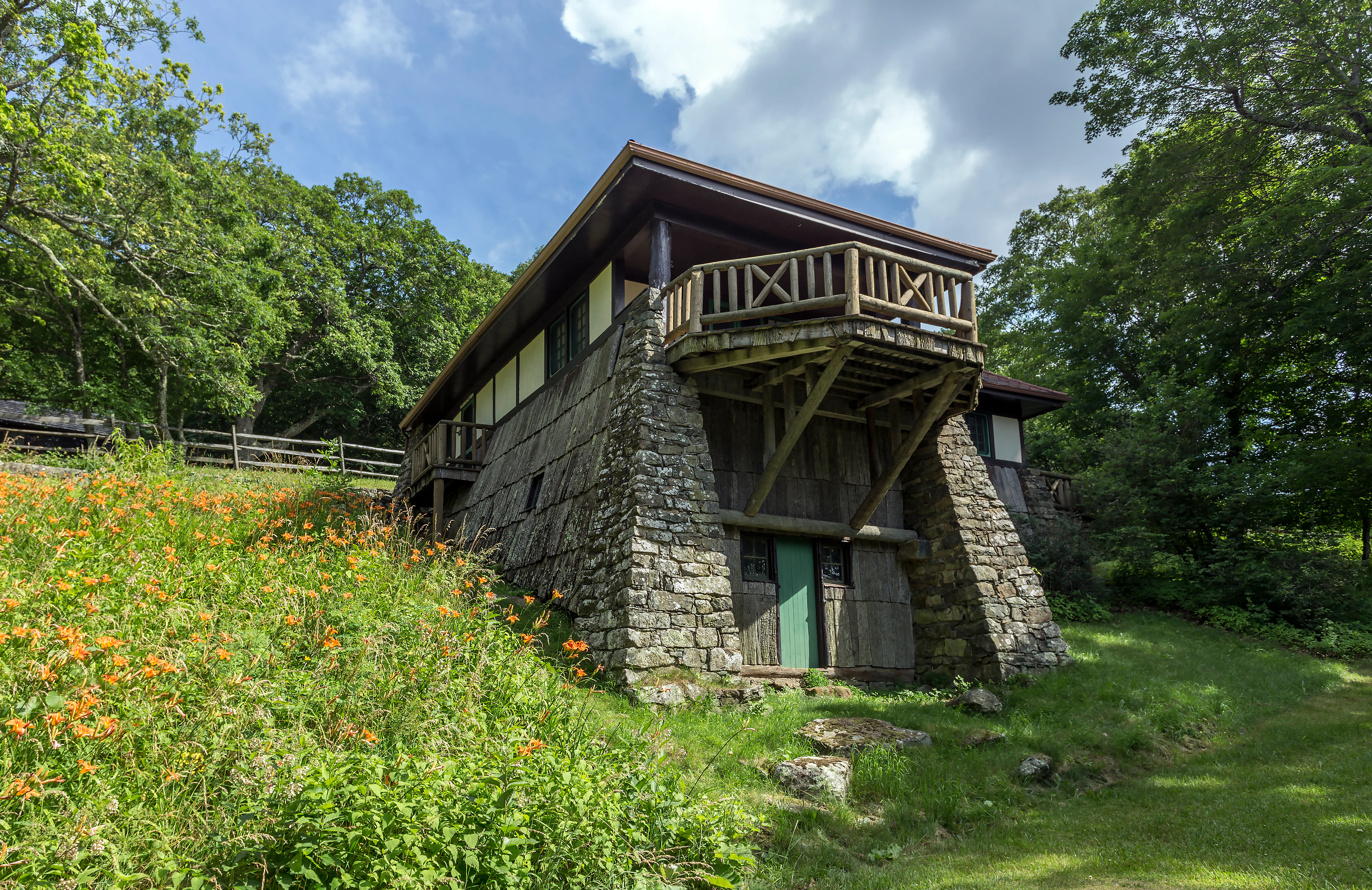 Stone and wood lodge building with a wooden balcony, surrounded by green grass and orange wildflowers, with trees in the background