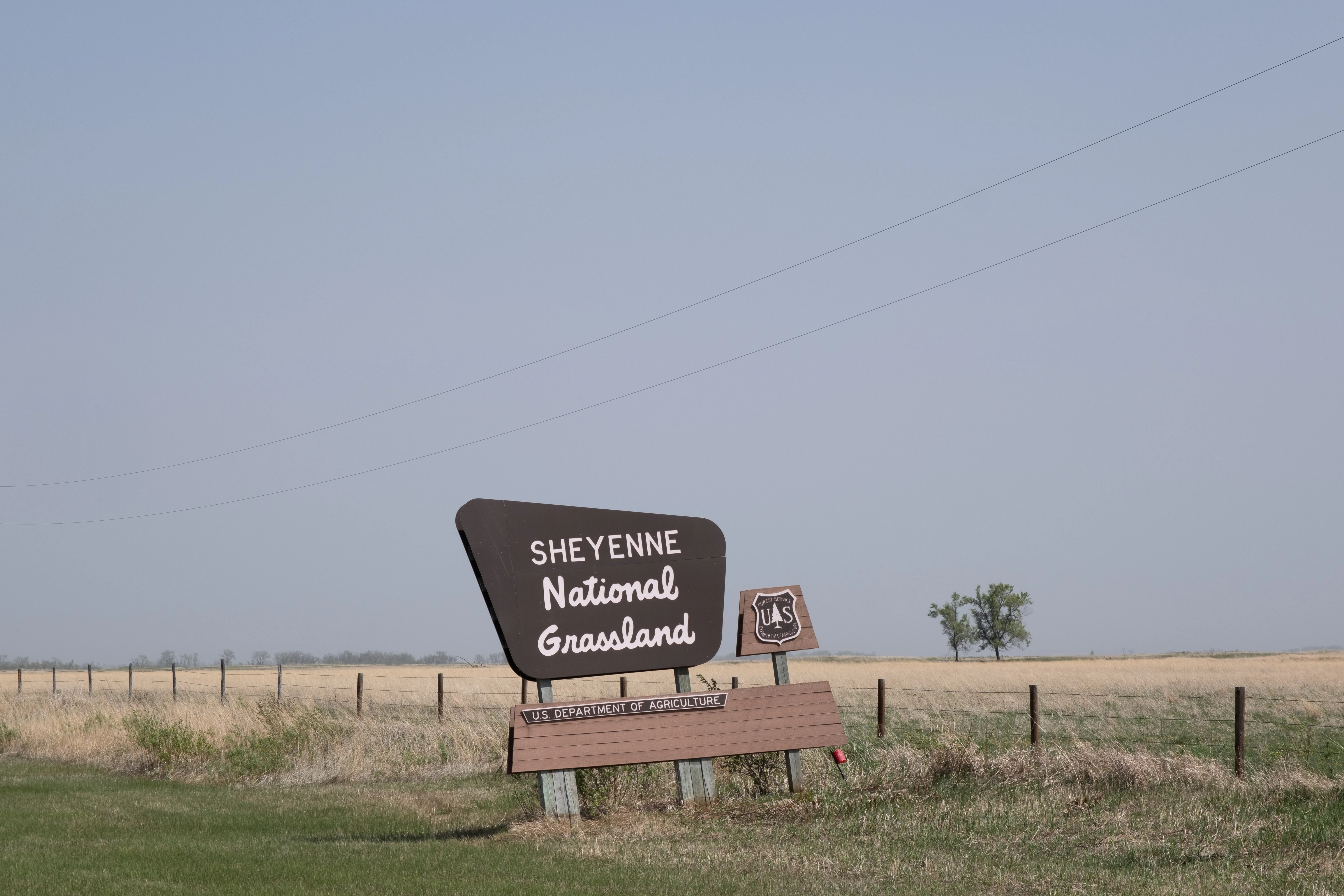 A brown road sign for Sheyenne National Grassland stands in a flat, open prairie landscape with a wire fence and a few distant trees.