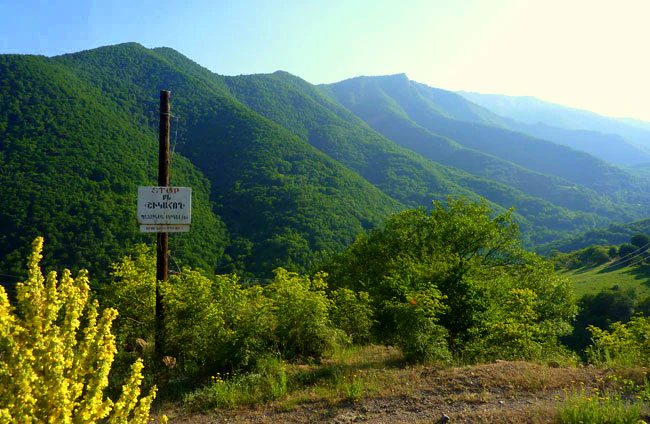 A panoramic landscape view of green, forested mountains under a bright sky, with a tall wooden post featuring a white sign in the foreground and yellow wildflowers to the left.
