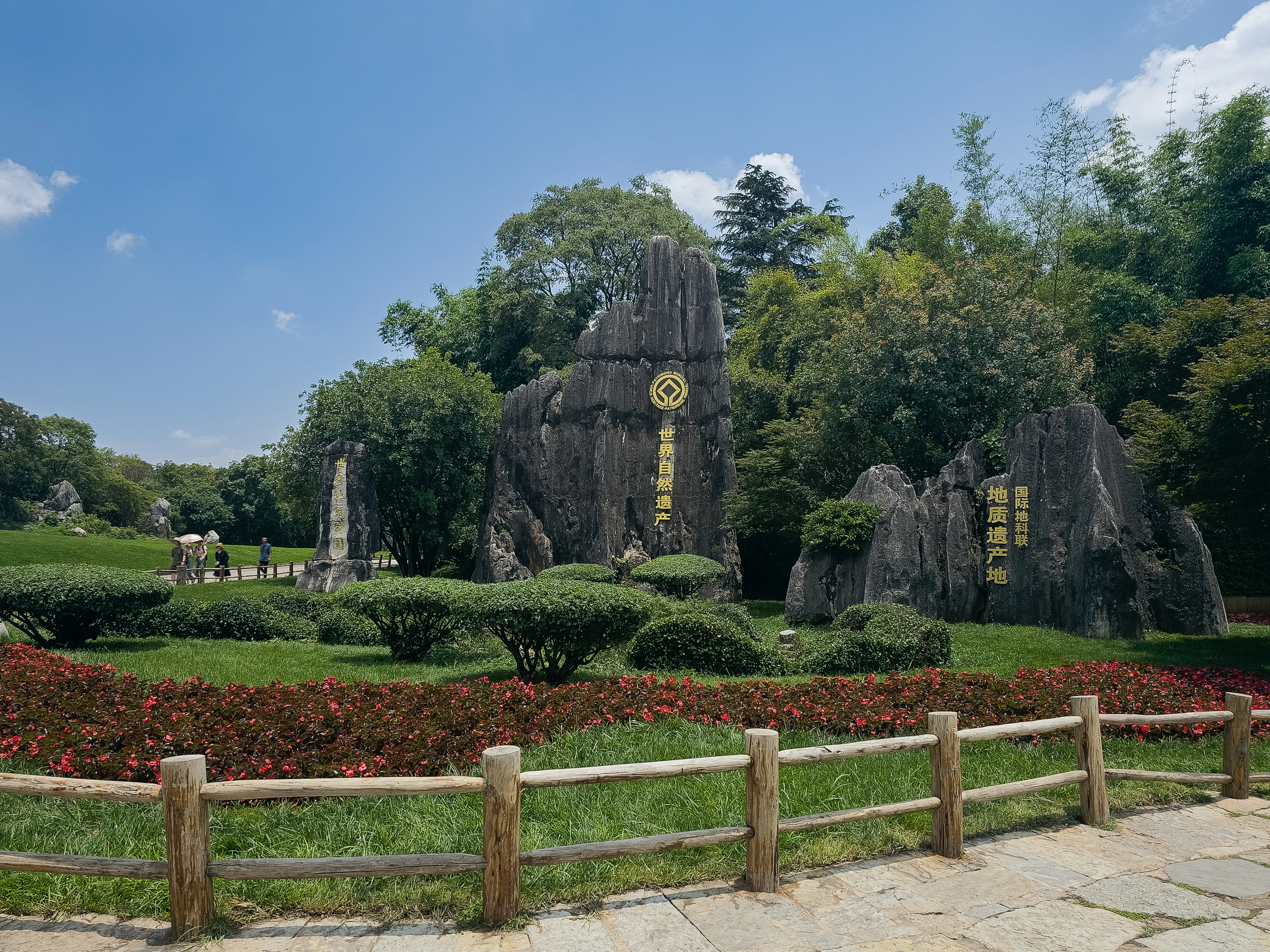Un jardín paisajista con parterres de flores rojas y una valla de madera en primer plano, setos verdes recortados en el plano medio y grandes formaciones de piedra caliza oscura con caracteres chinos dorados y un emblema circular al fondo bajo un cielo azul.