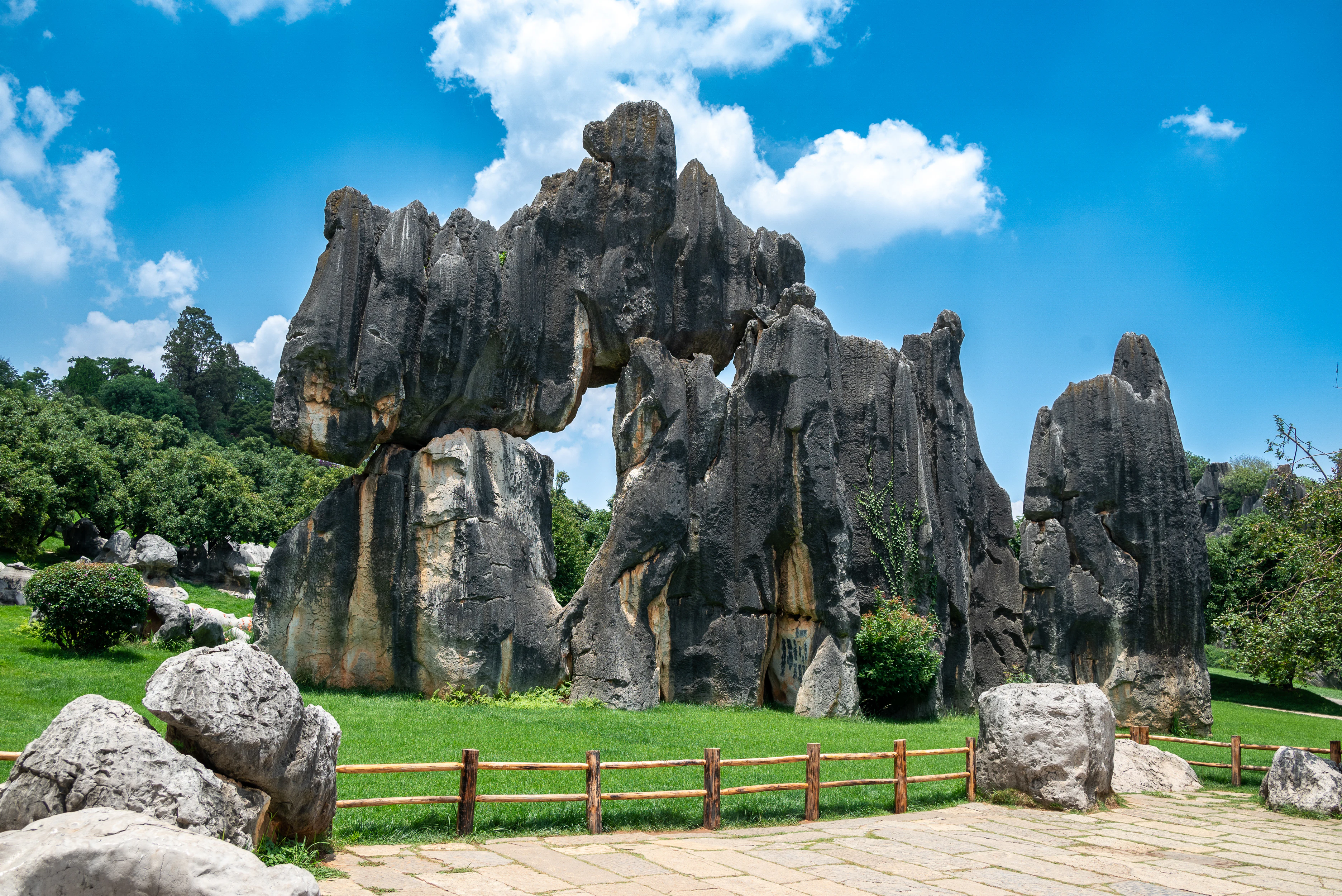 Una vista de grandes formaciones rocosas de karst de color gris oscuro en un bosque de piedra, rodeadas de hierba verde, una valla de madera y un sendero pavimentado bajo un cielo azul con nubes blancas.