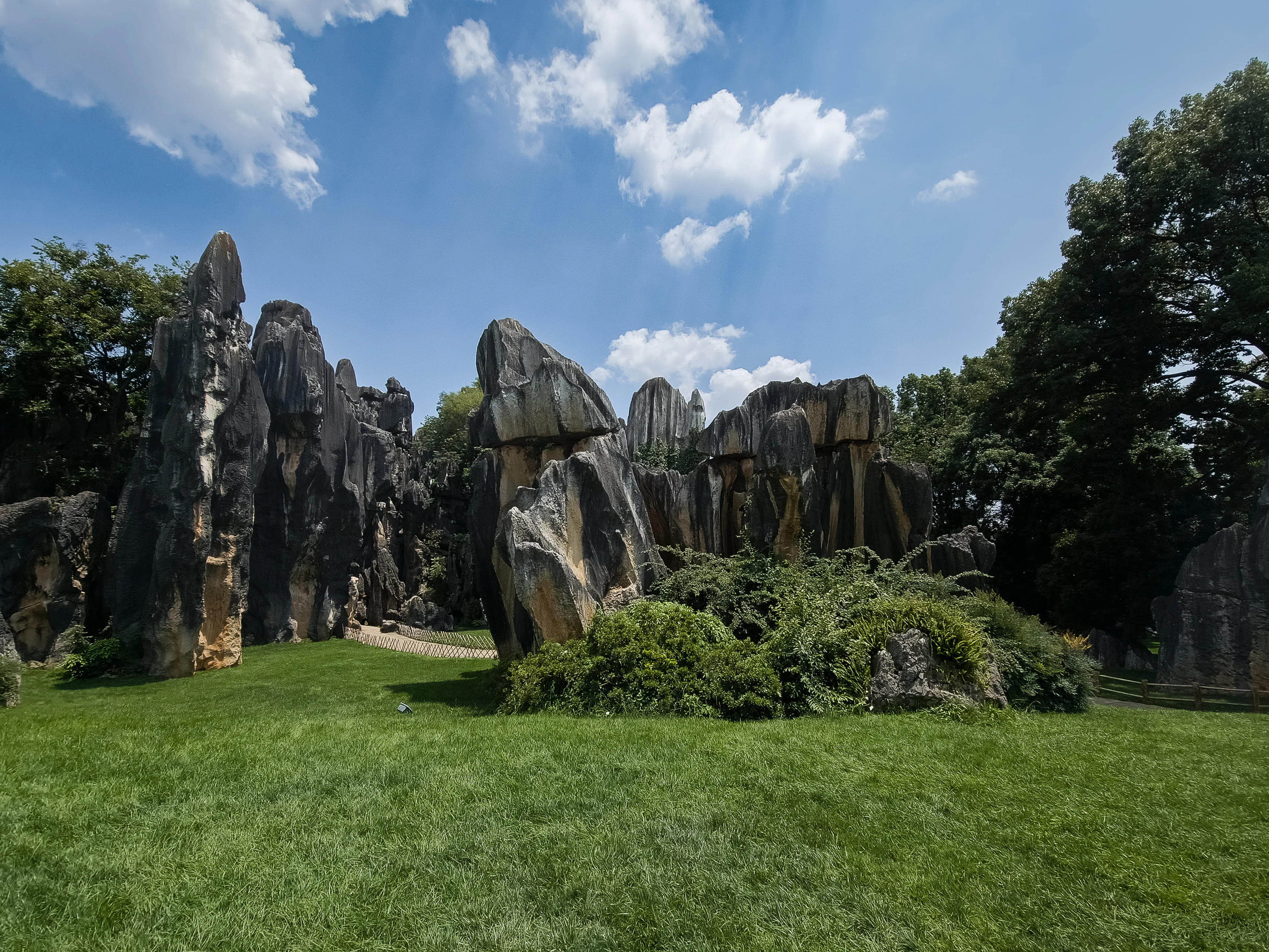 Altas formaciones rocosas de piedra caliza, de tonos grises y marrones, que se alzan sobre un campo de hierba verde con árboles al fondo, bajo un cielo azul con nubes blancas.