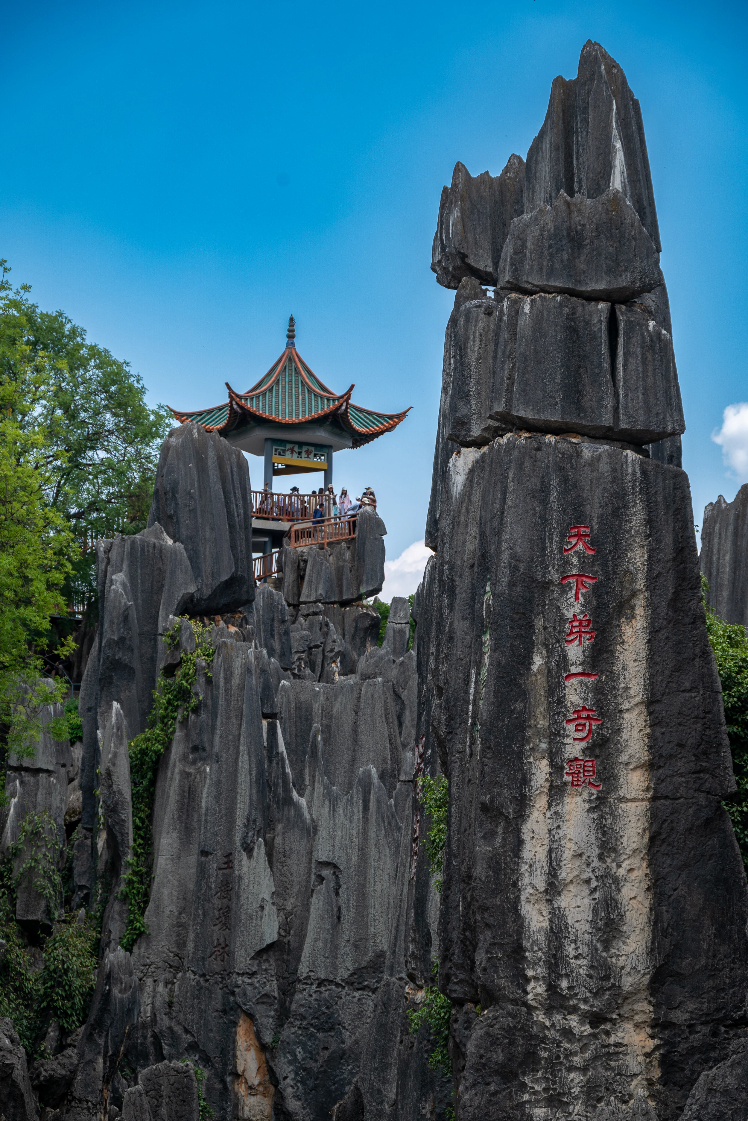 Vista vertical de formaciones rocosas de piedra caliza gris oscuro que se alzan contra un cielo azul. Un pabellón tradicional chino con un techo curvo y una plataforma de observación se encuentra entre las rocas en el plano medio, con varias personas de pie sobre él. Grandes caracteres chinos rojos están pintados verticalmente en la formación rocosa prominente en el primer plano derecho. Árboles y vegetación verde crecen alrededor de la base y entre las rocas.