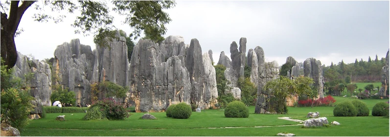 Vista panorámica de formaciones rocosas de piedra caliza gris, tipo karst, que se alzan sobre un césped verde con arbustos y árboles recortados de forma redondeada, bajo un cielo nublado, con algunas figuras pequeñas visibles cerca de la base de las rocas.