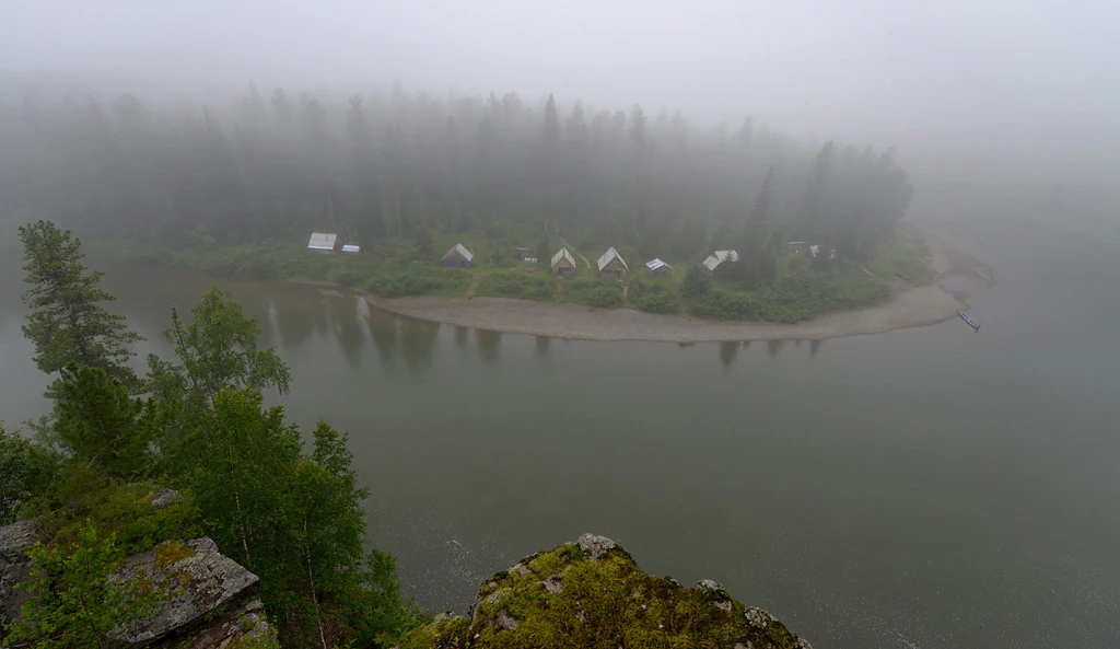 Una vista gran angular de un río que serpentea alrededor de una orilla boscosa donde se ubican una serie de pequeñas cabañas o puestos avanzados, todo envuelto en una densa niebla matinal.