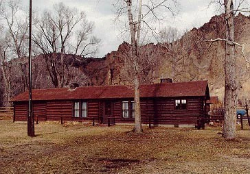 A long, single-story dark brown log cabin building stands on a flat clearing, backed by tall, craggy, light-brown cliffs and leafless trees.