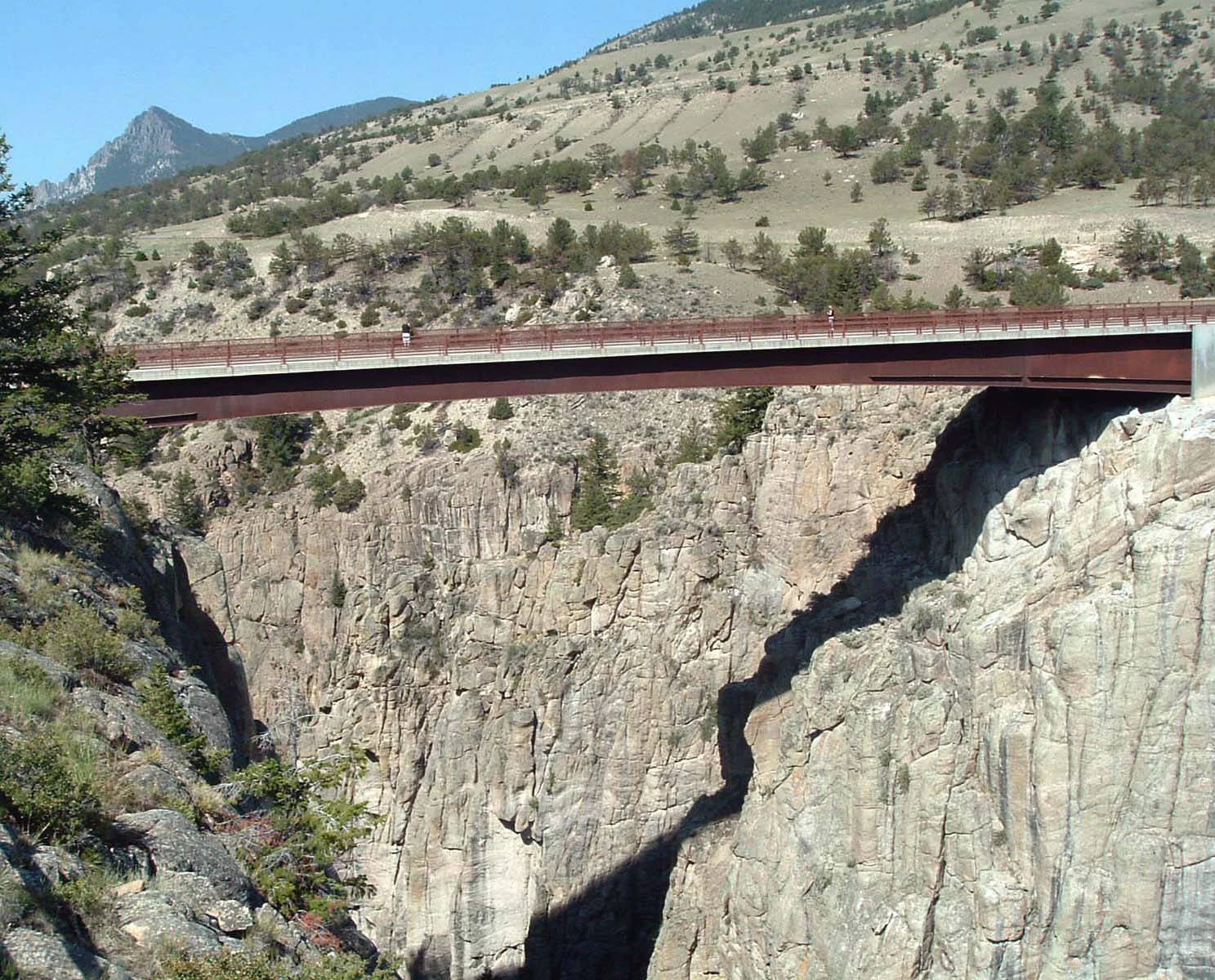 A steel girder bridge spans a deep, rugged canyon with vertical rocky walls, surrounded by mountain slopes with scattered coniferous trees.