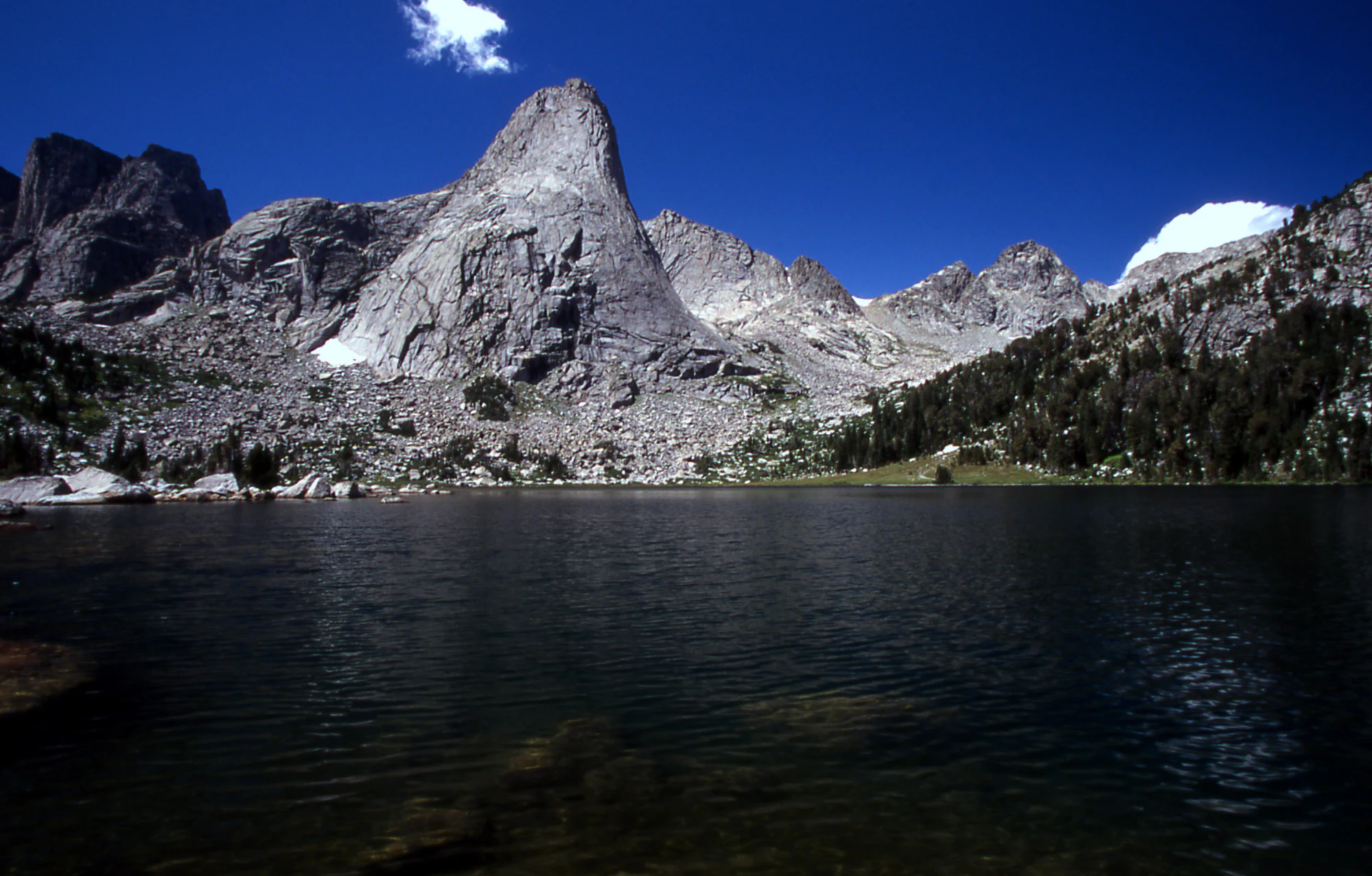 A towering granite mountain peak reflected in the calm waters of a high alpine lake, with coniferous forest on the right shoreline.