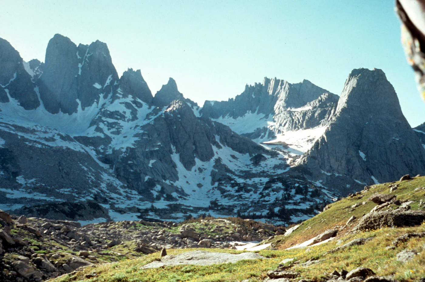 A wide landscape view of jagged granite mountain peaks with patches of snow, overlooking a rocky valley with green meadow and sparse vegetation.