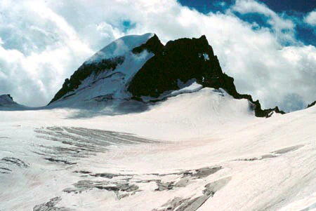A jagged, dark mountain peak rises above a vast, expansive snow and ice field under a cloudy sky.
