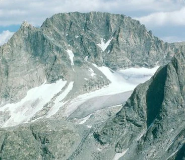 A wide, eye level shot of the craggy, grey, snow-dusted peak of Gannett Peak with a glacier nestled in the mountain cirque.