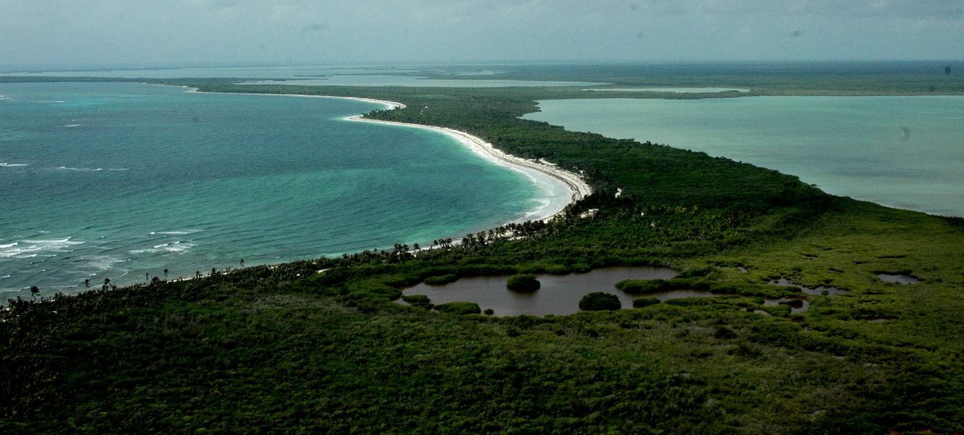 Aerial perspective of a narrow peninsula separating the Caribbean Sea from a coastal lagoon, featuring dense green vegetation and a white sand beach.