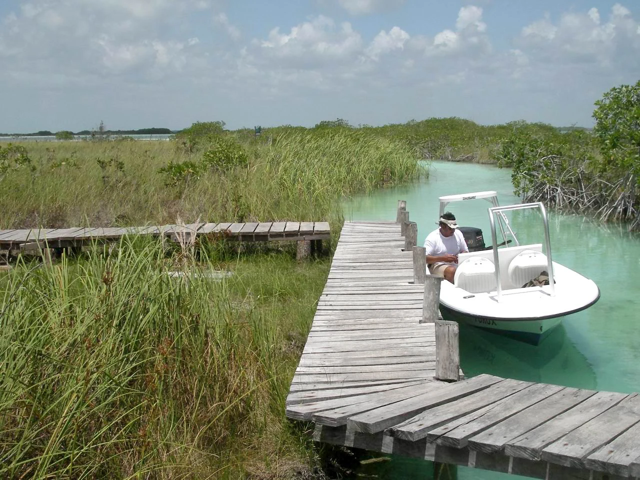 A small white motorboat is docked at a weathered wooden boardwalk that extends through a grassy marshland with clear light turquoise water.