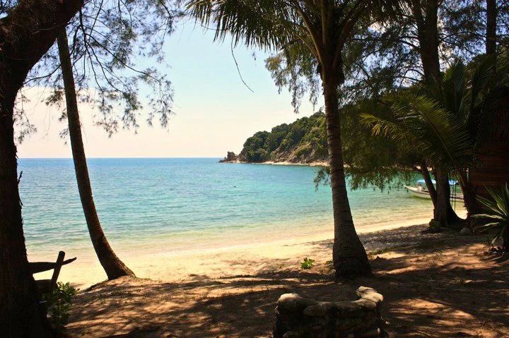 A scenic view from a shaded, tree-lined beach looking out over a turquoise bay towards a forested island headland in Malaysia.