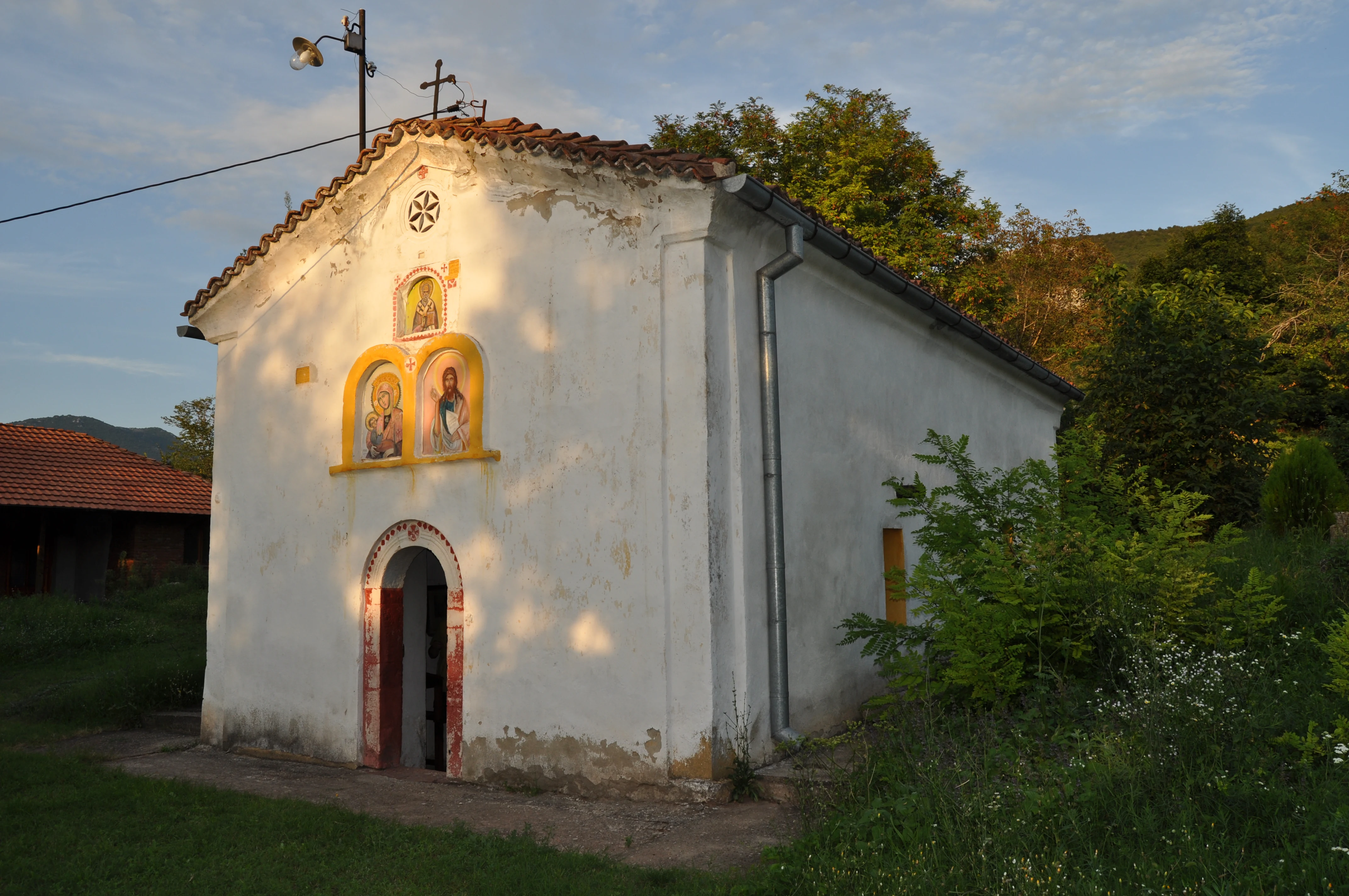 White church with yellow-framed religious icons above the entrance, surrounded by greenery and hills