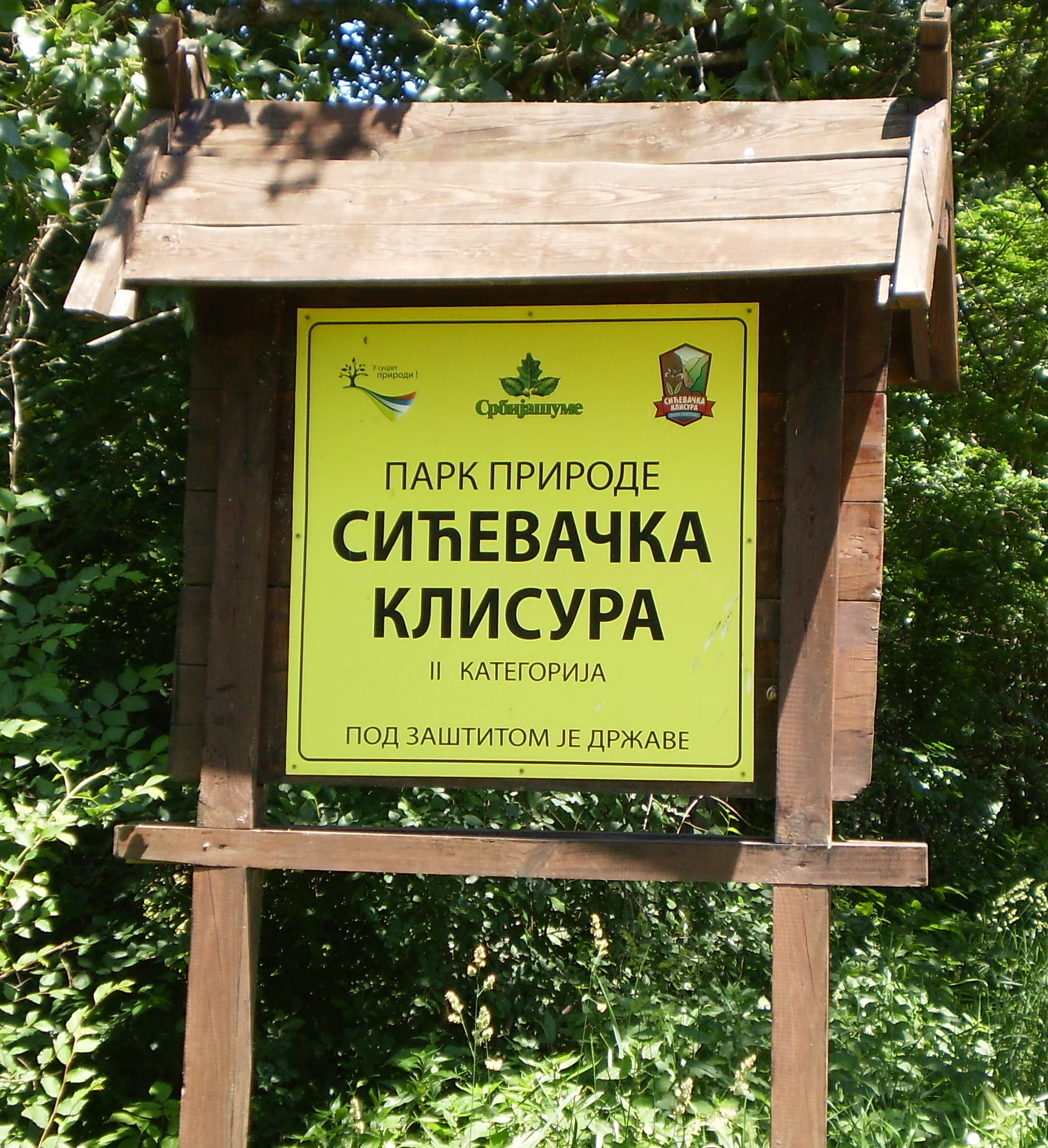 Yellow sign with Cyrillic text 'ПАРК ПРИРОДЕ СИЋЕВАЧКА КЛИСУРА' mounted on wooden structure, surrounded by green foliage
