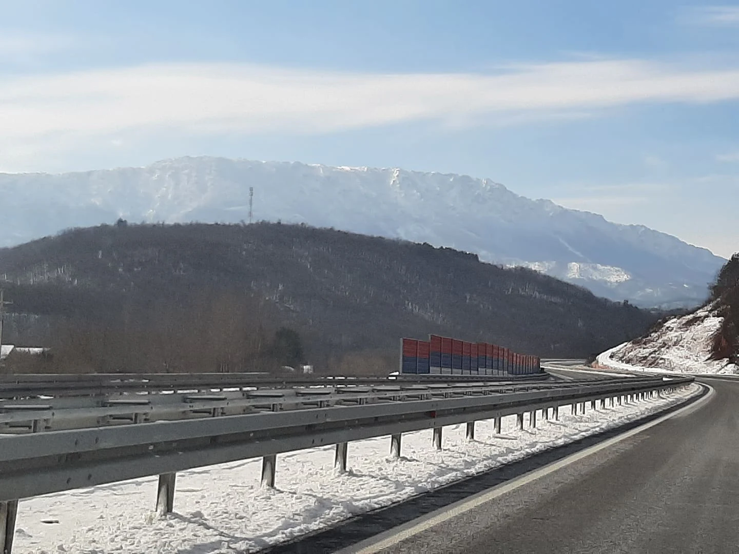 Highway with guardrails running through a mountainous landscape with snow-covered ground and distant mountain peaks under a clear sky