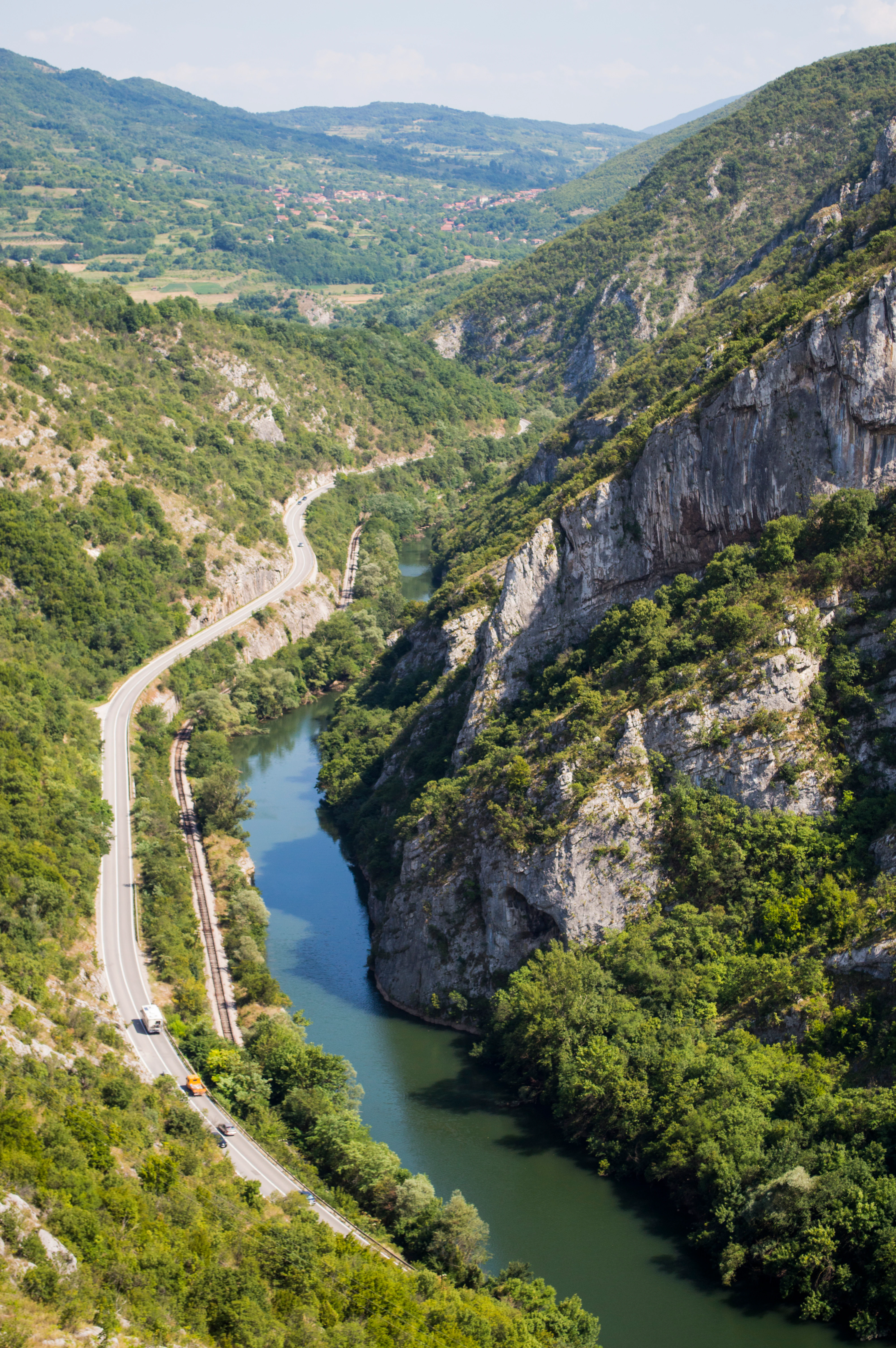 Aerial view of a river flowing through a gorge with steep limestone cliffs, a road with vehicles, and forested slopes