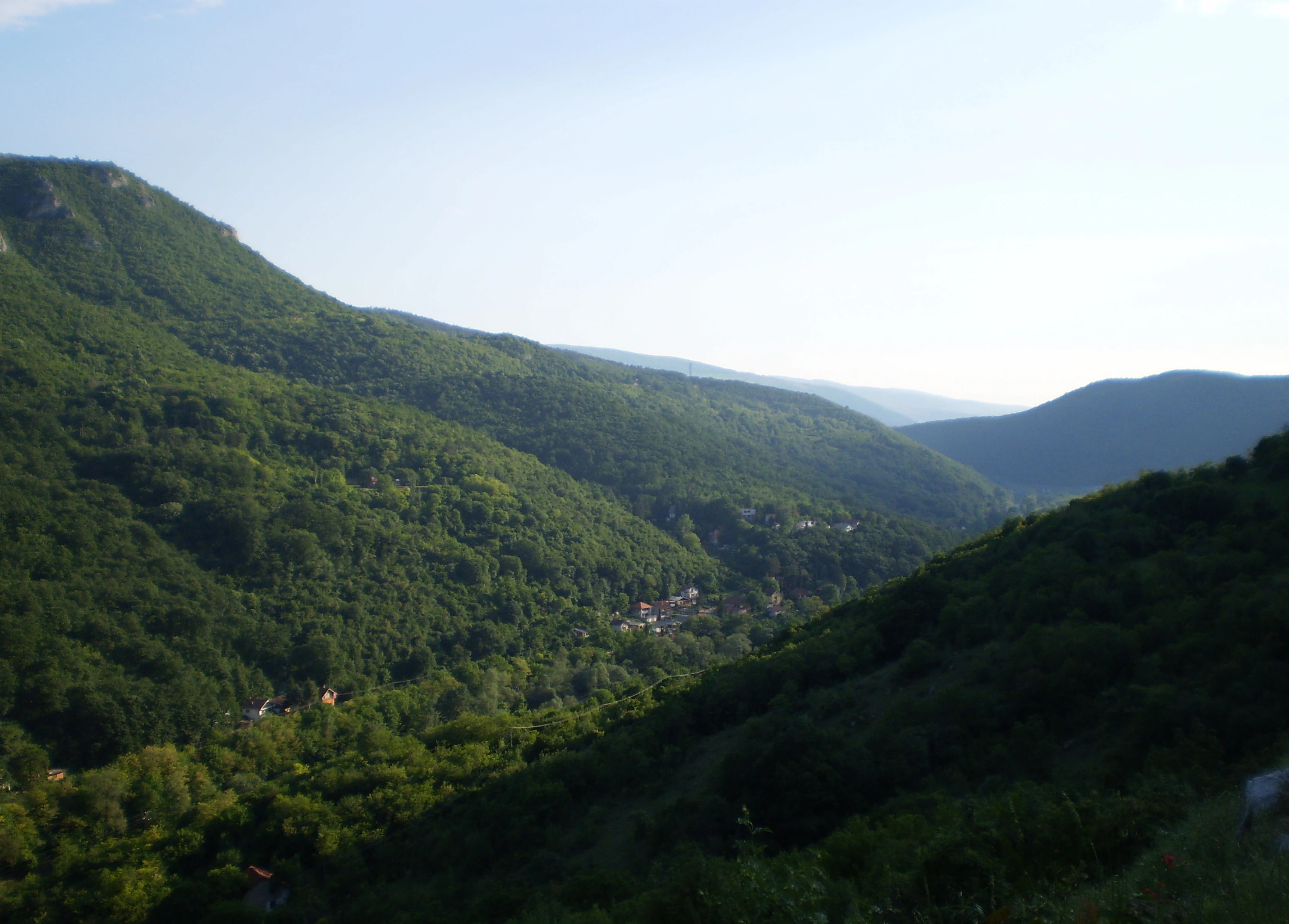 Wide landscape view of a forested valley with rolling hills and small settlements in the distance under a clear sky