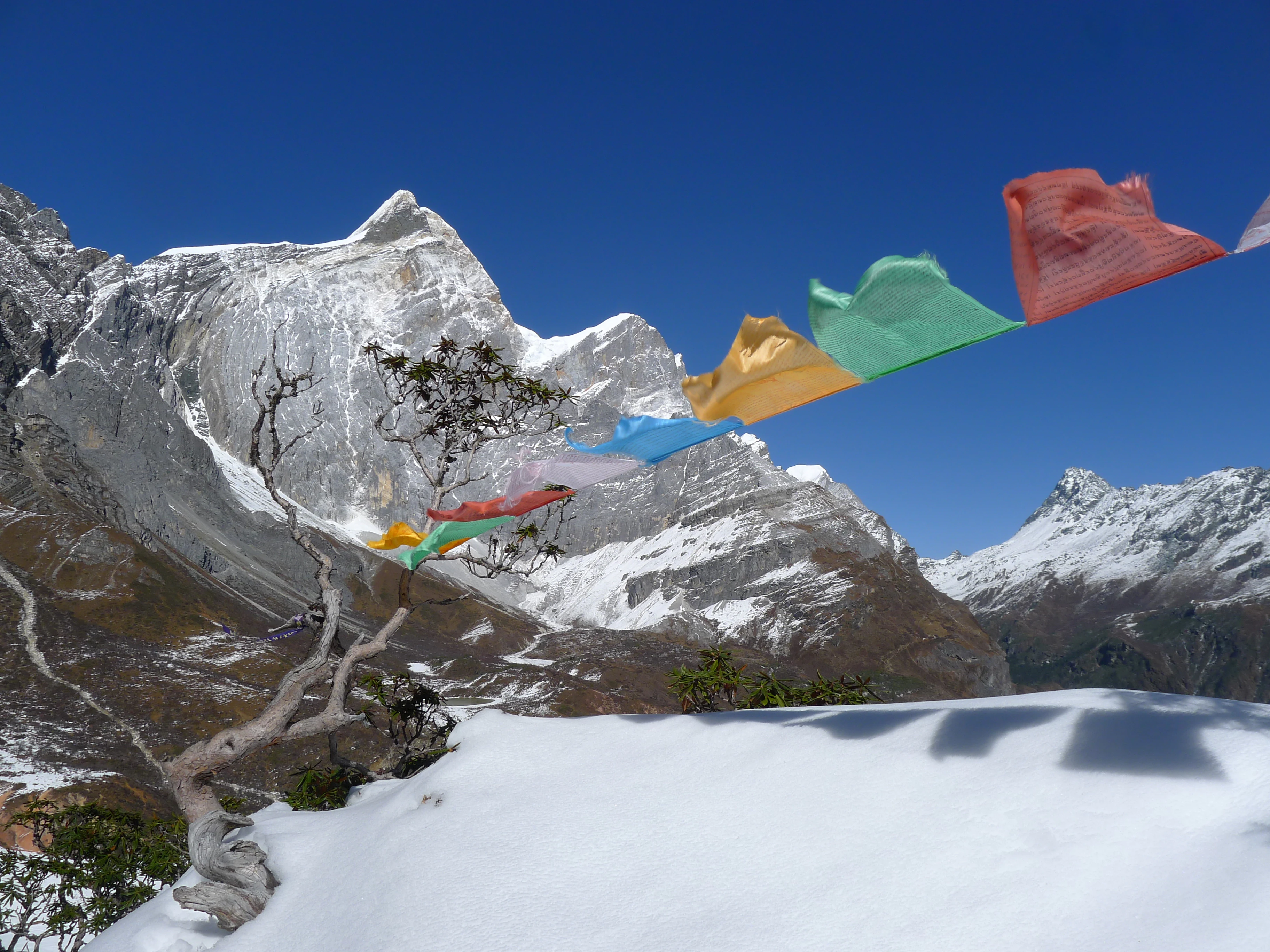A line of colorful Tibetan prayer flags stretches across the foreground, framing a sharp, snow-covered mountain peak against a deep blue sky.