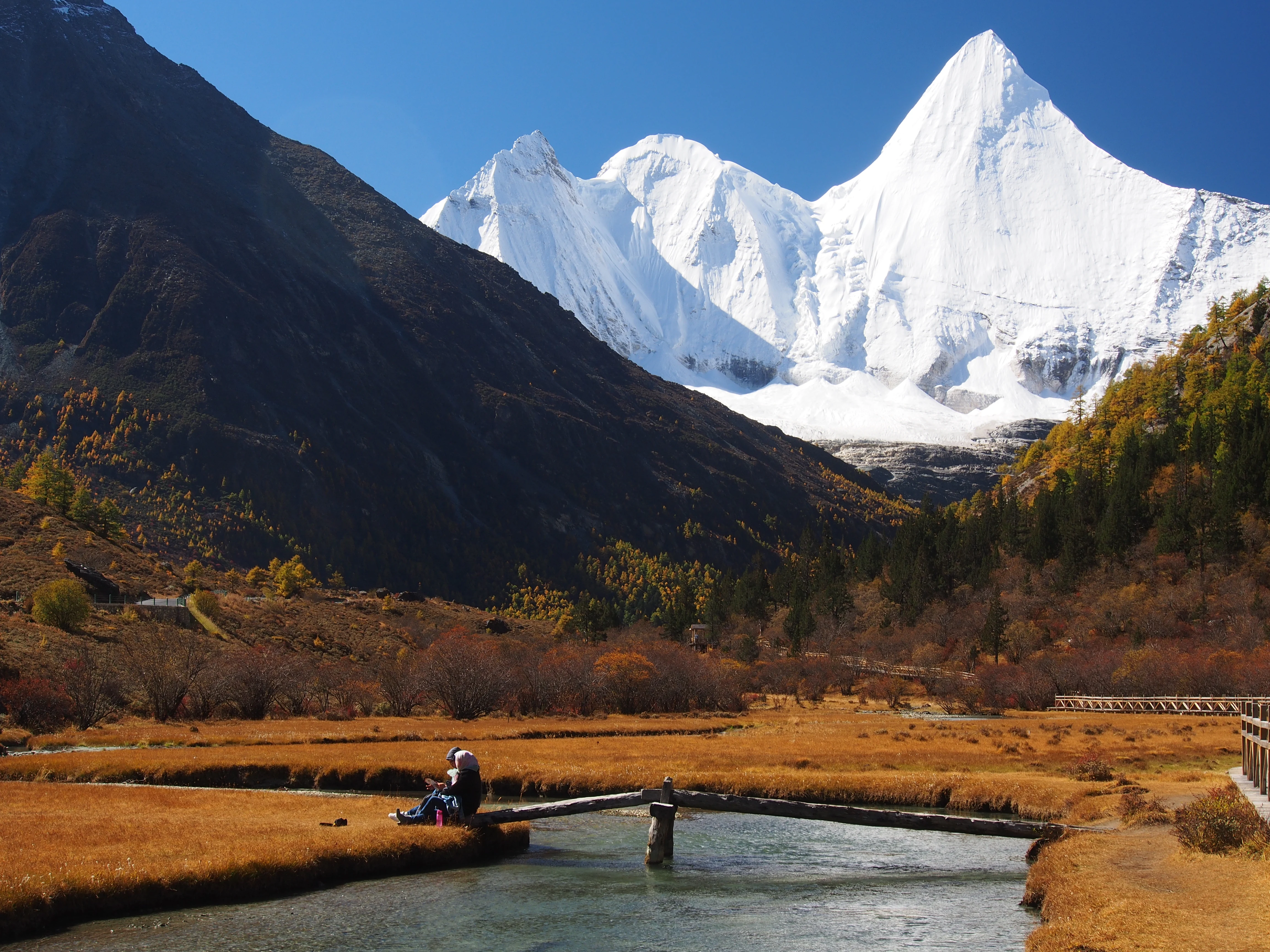 A snowy, pointed mountain peak rises above a golden-brown alpine meadow and a clear, flowing river, with a wooden bridge in the foreground.