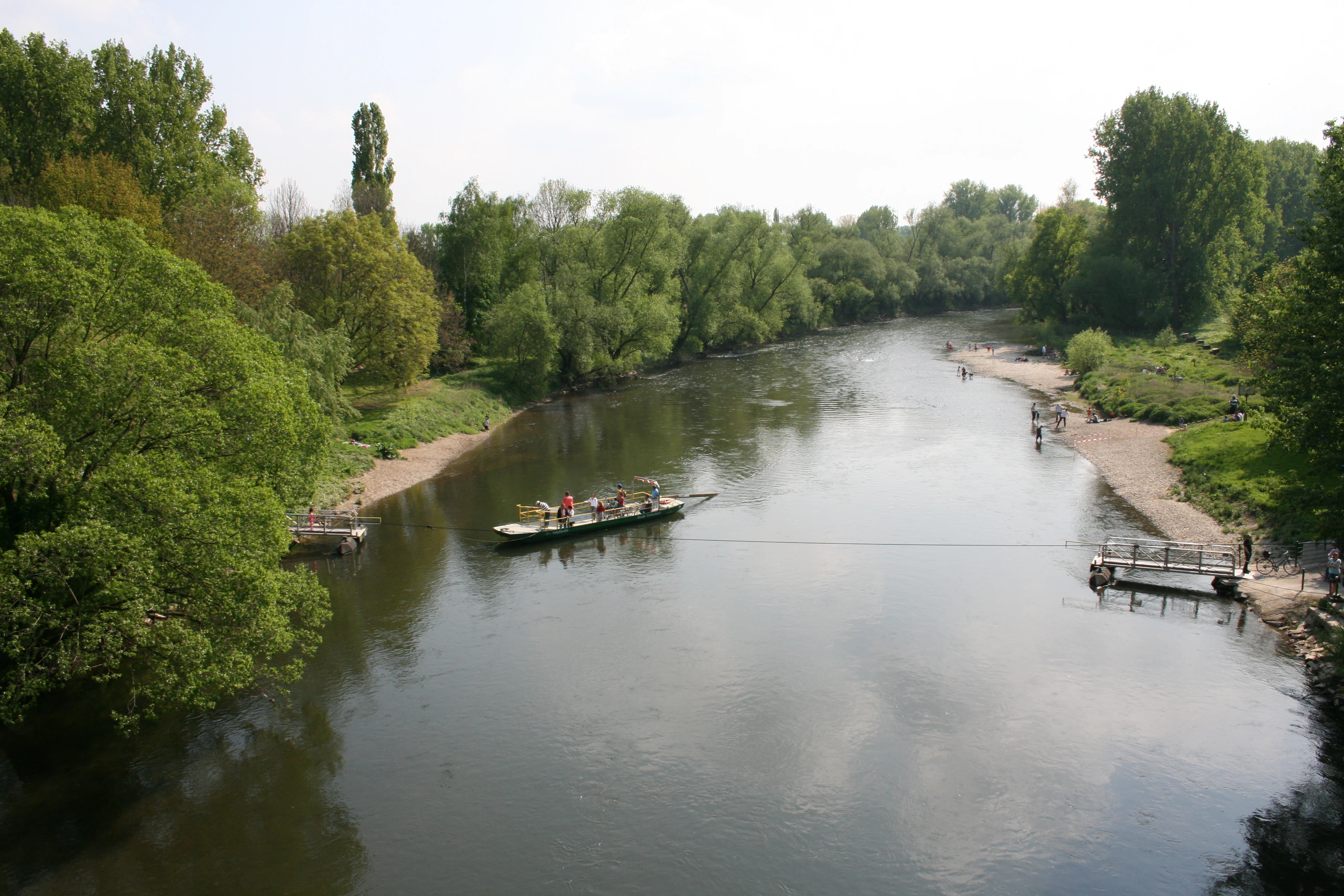A small reaction ferry tethered to a cable crossing the slow moving Sieg River, surrounded by lush green floodplain trees and sandy banks.