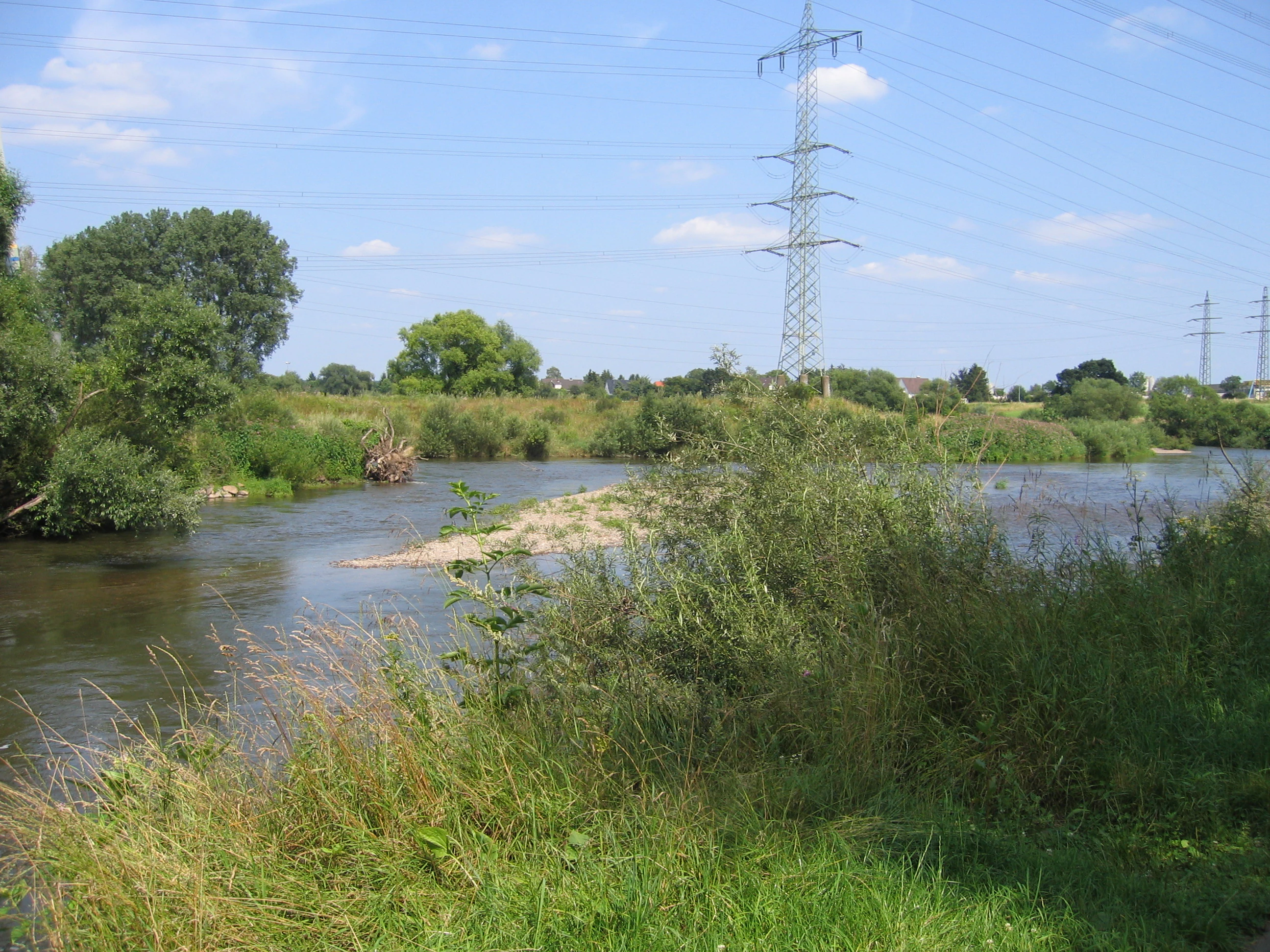 A wide view of the Sieg river with a small gravel island in the center, surrounded by green riparian vegetation and bushes, under a clear blue sky with a tall metal electrical pylon in the background.