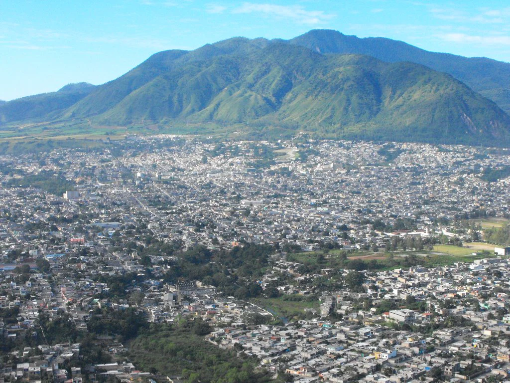 A high-angle view showing a sprawling city in the foreground, leading up to a large, green, rounded mountain range under a clear sky.