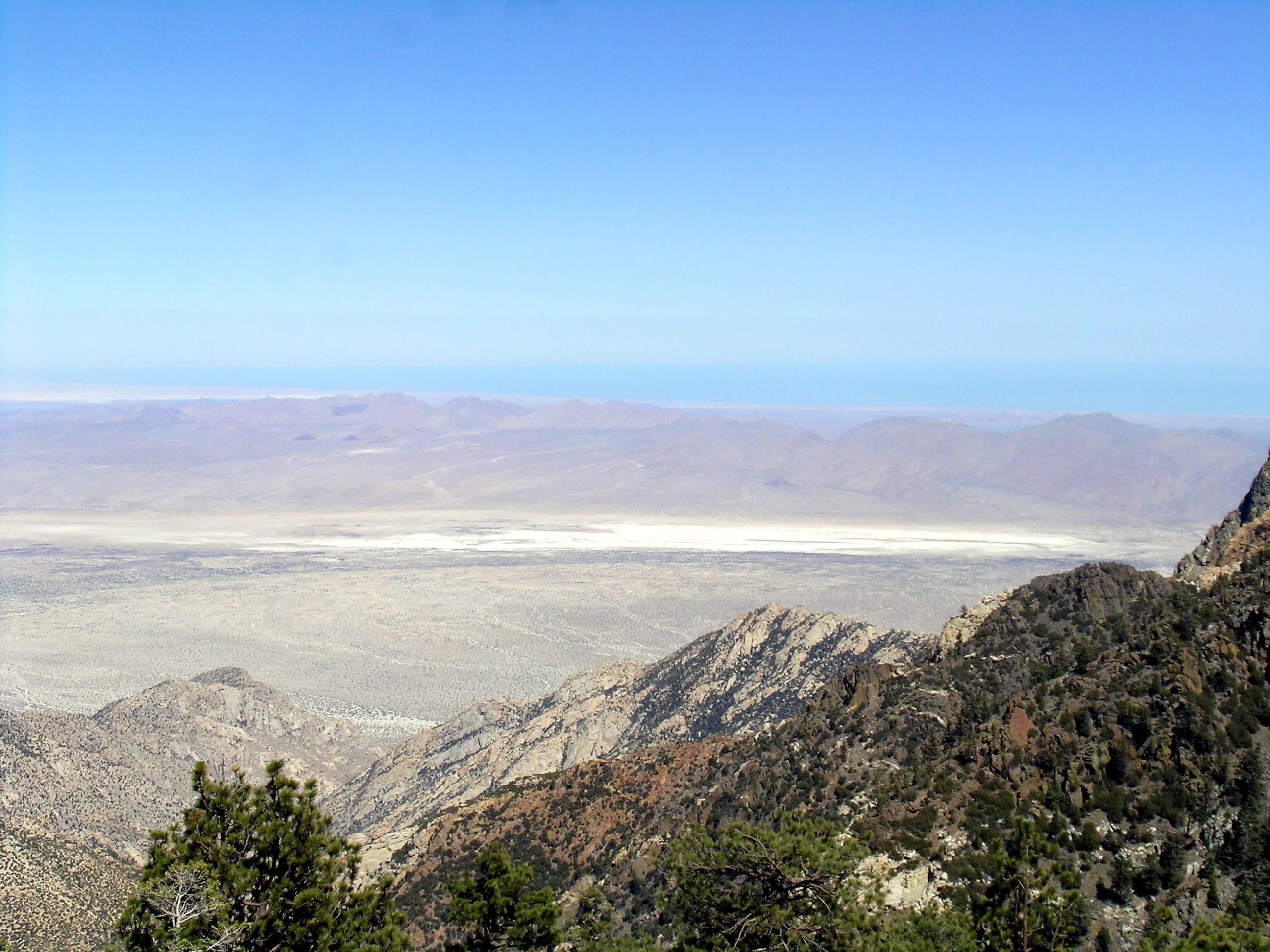 Mountain landscape with pine trees and rocky slopes in foreground, extending to a valley and distant mountain ranges under clear blue sky