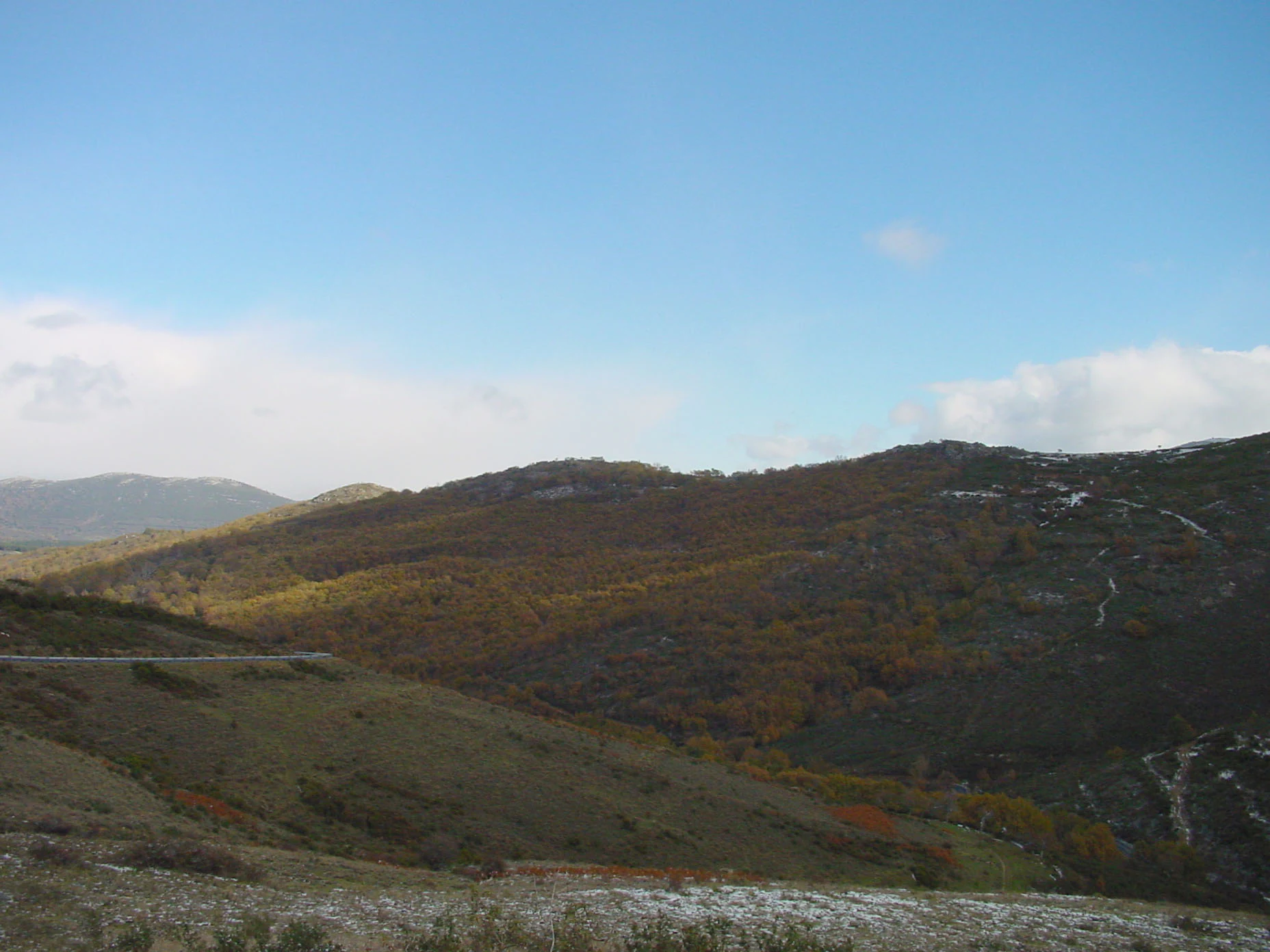 A wide landscape view of rolling hills and forested slopes under a clear blue sky, with a dusting of snow in the foreground.