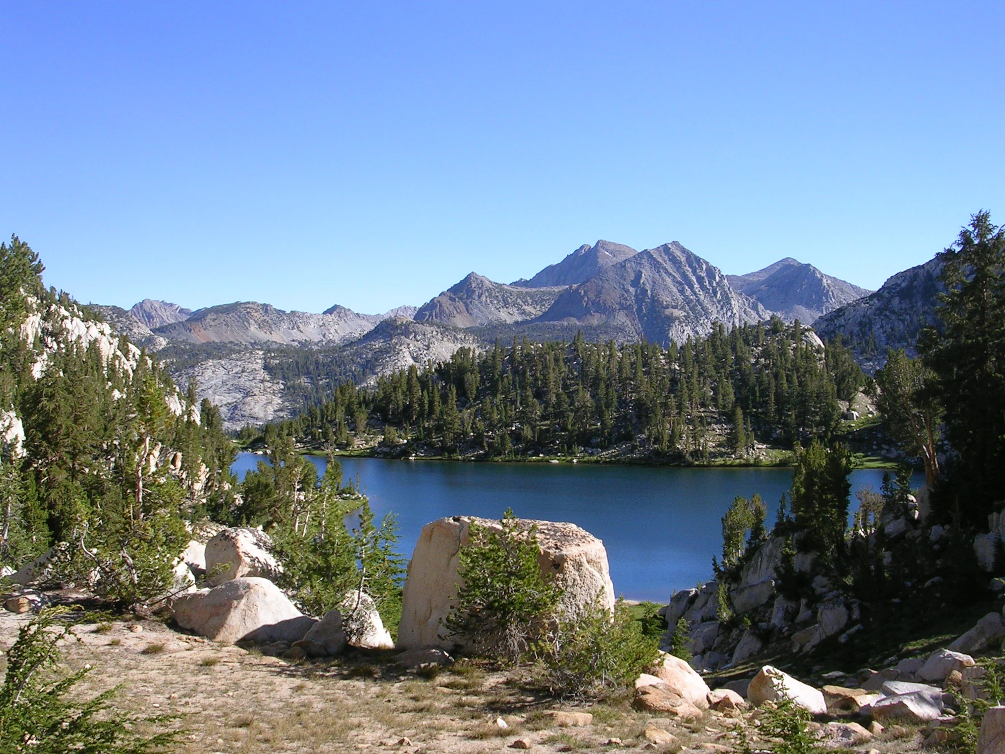 A clear blue alpine lake surrounded by coniferous trees and granite peaks under a cloudless sky.