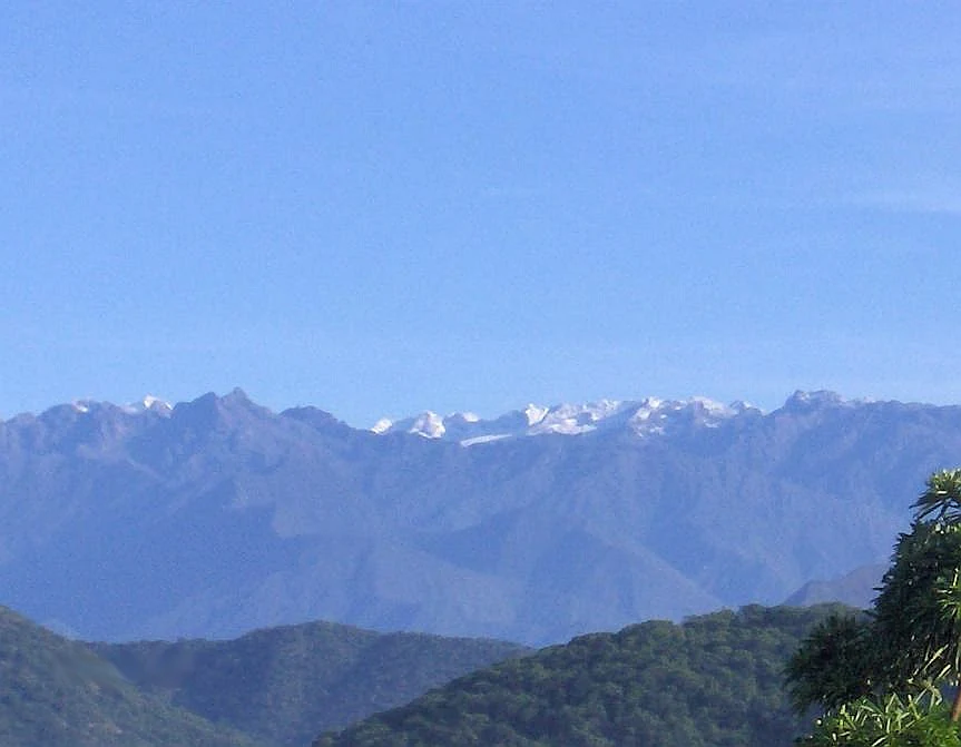 Cielo azul claro sobre cordillera con picos nevados a lo lejos y colinas boscosas en primer plano