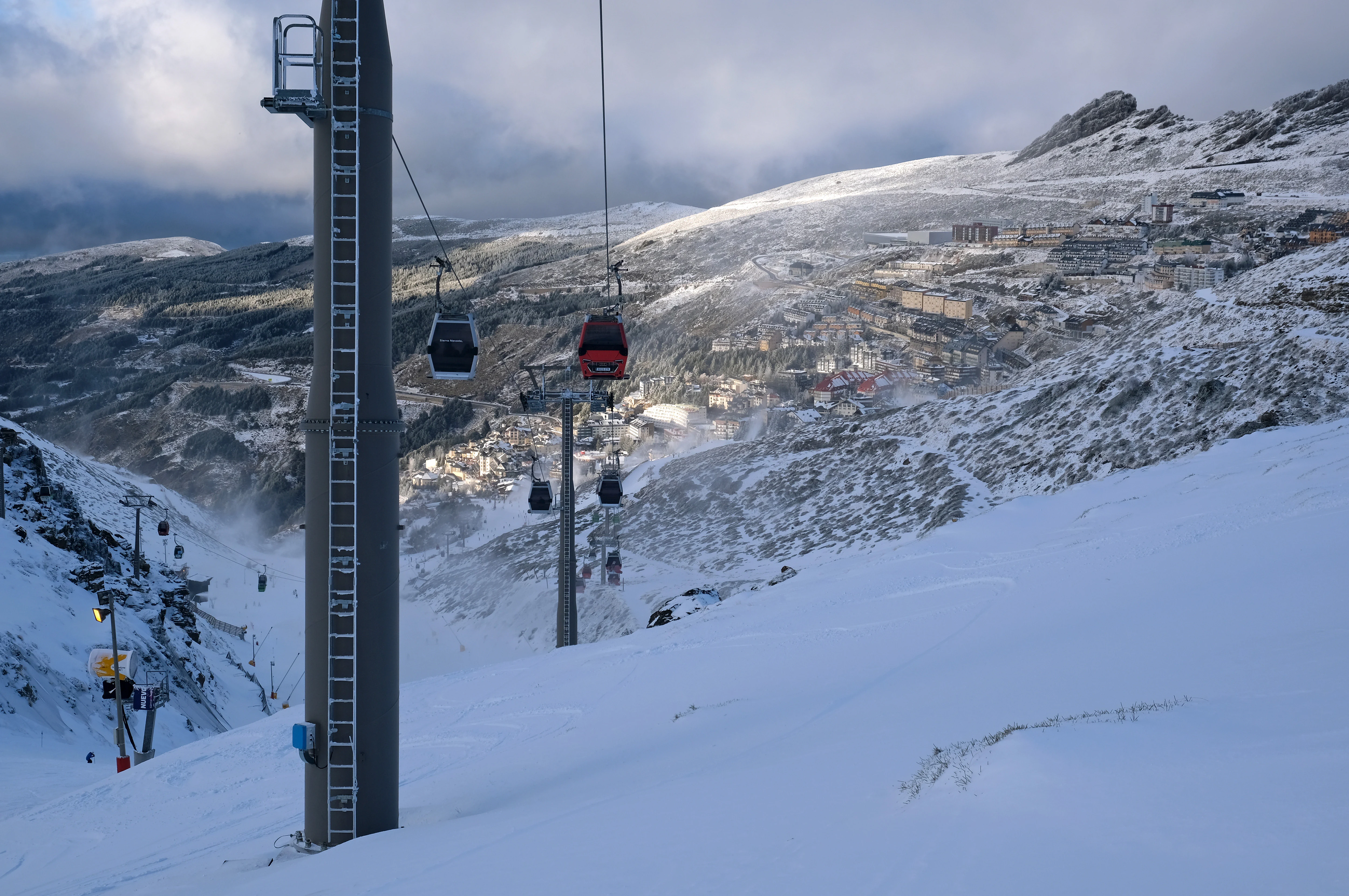 Ski lift towers with red and blue gondolas on a snowy mountain slope with valley and distant mountains under cloudy sky