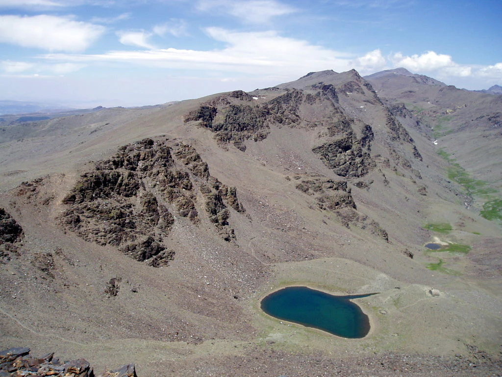 A high-altitude lake surrounded by rocky mountain slopes under a partly cloudy sky.
