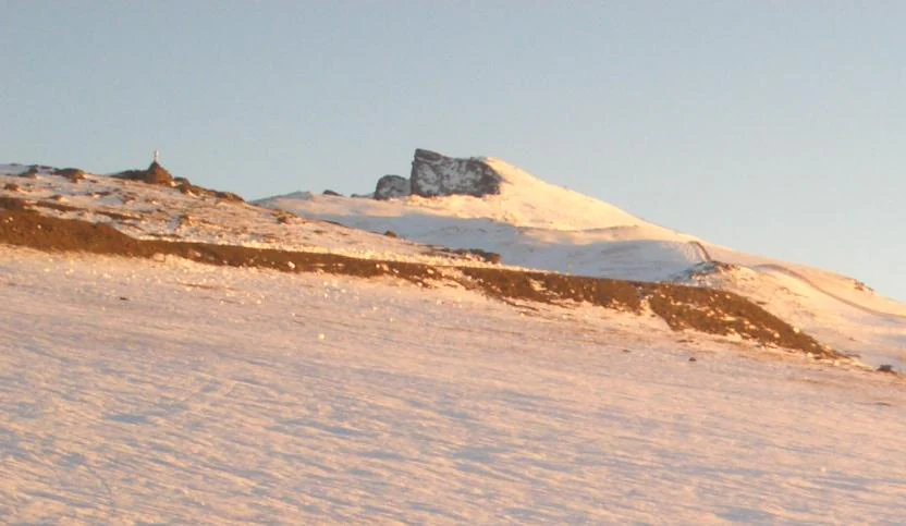 Snow-covered mountain peak with rocky outcrops and a small structure visible on the left slope under a clear sky.