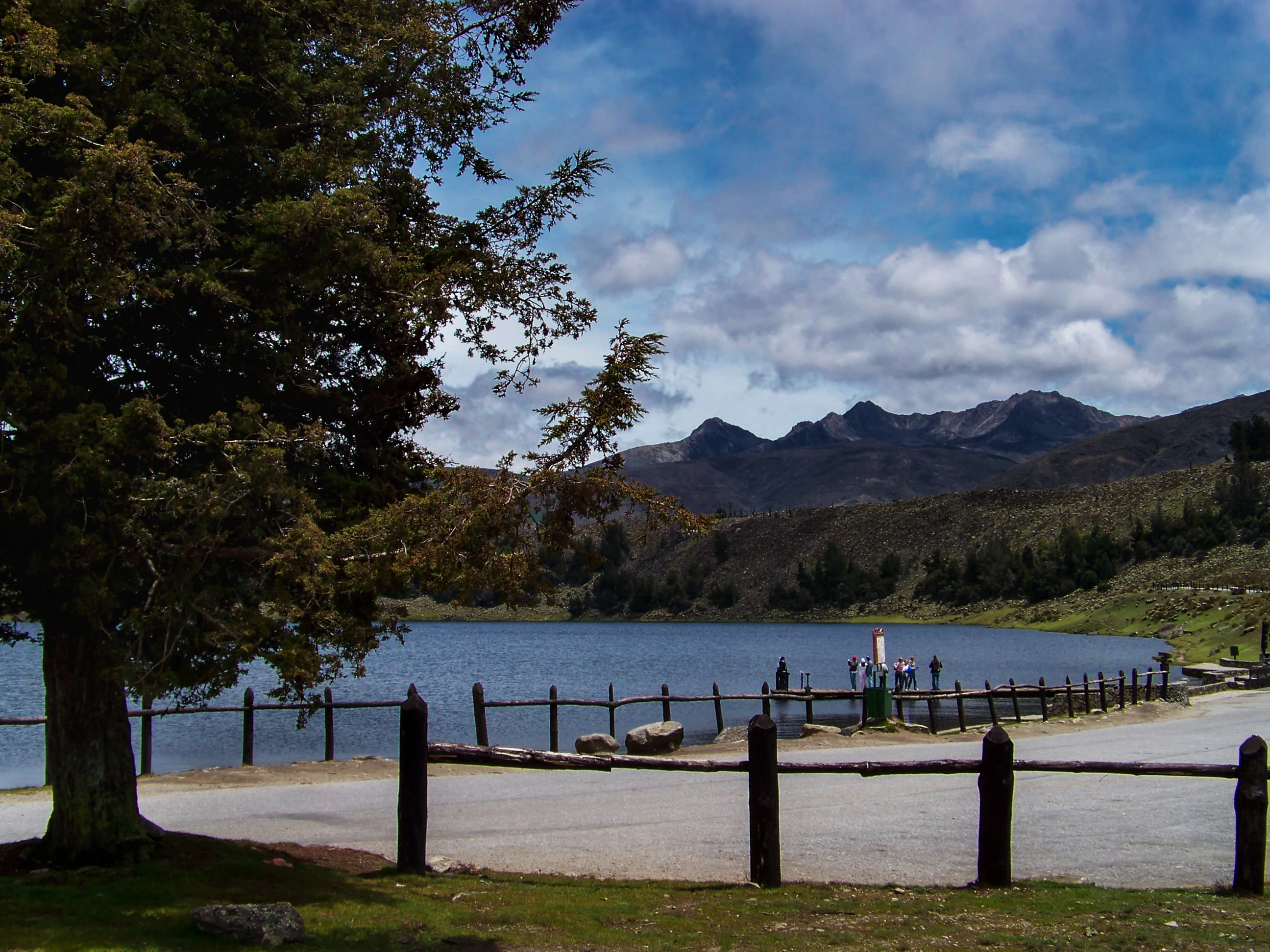 Un lago con montañas al fondo, una valla de madera en primer plano y personas reunidas en una zona de observación bajo un cielo parcialmente nublado