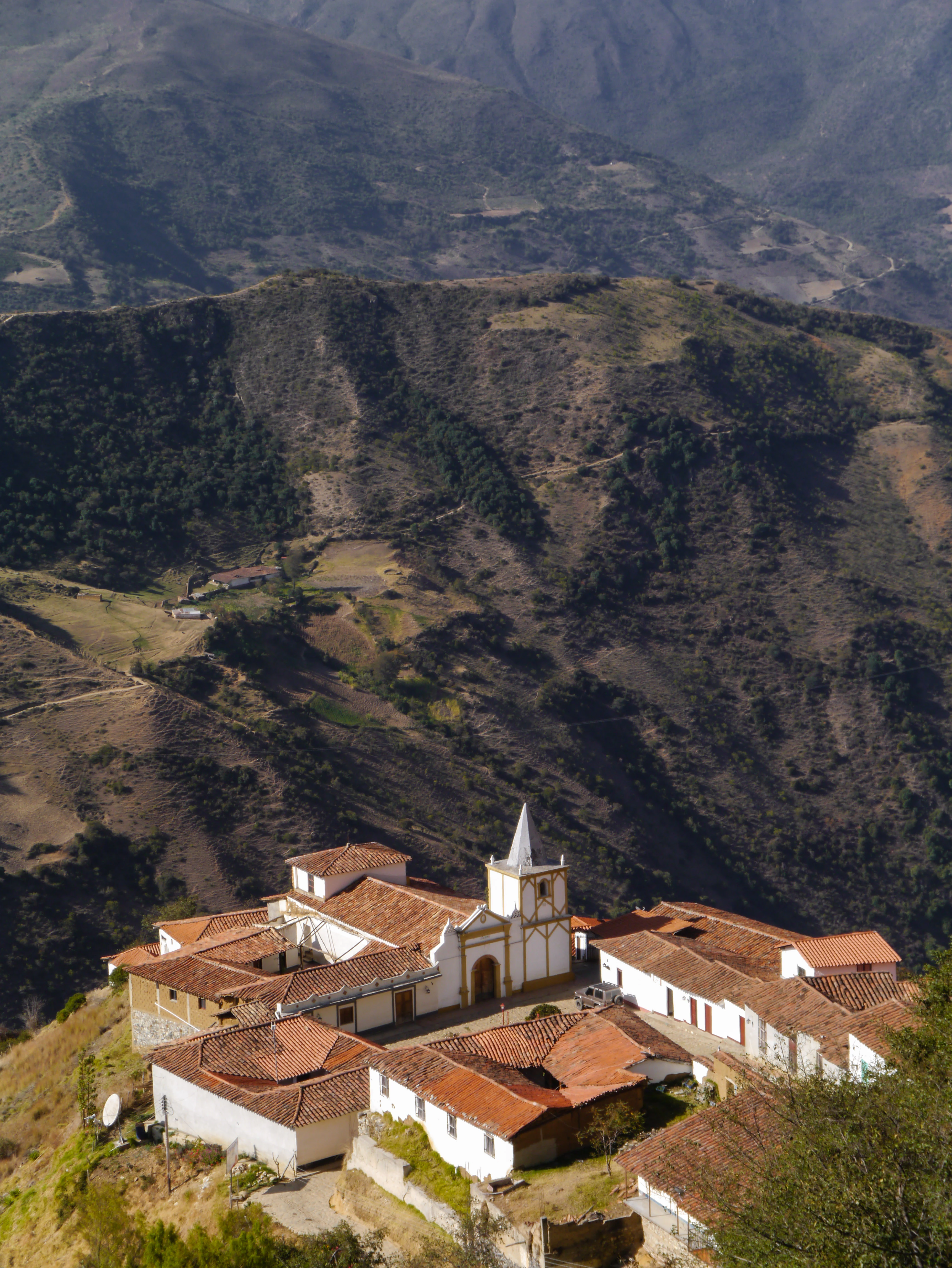 Un pueblo con edificios blancos y tejados de teja roja en una ladera con terreno montañoso al fondo