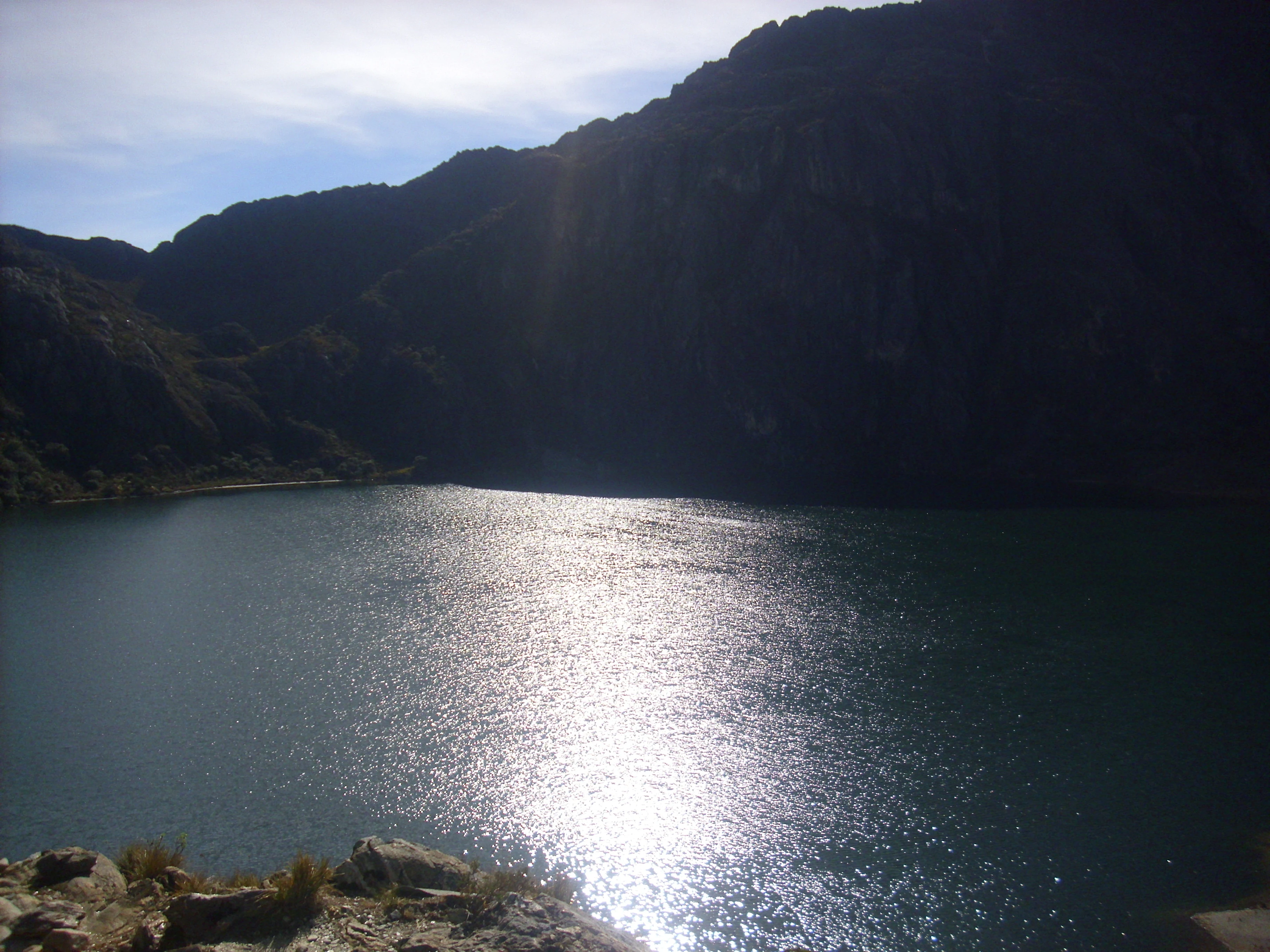 Lago iluminado por el sol que refleja la luz solar con montañas escarpadas y orilla rocosa bajo un cielo parcialmente nublado