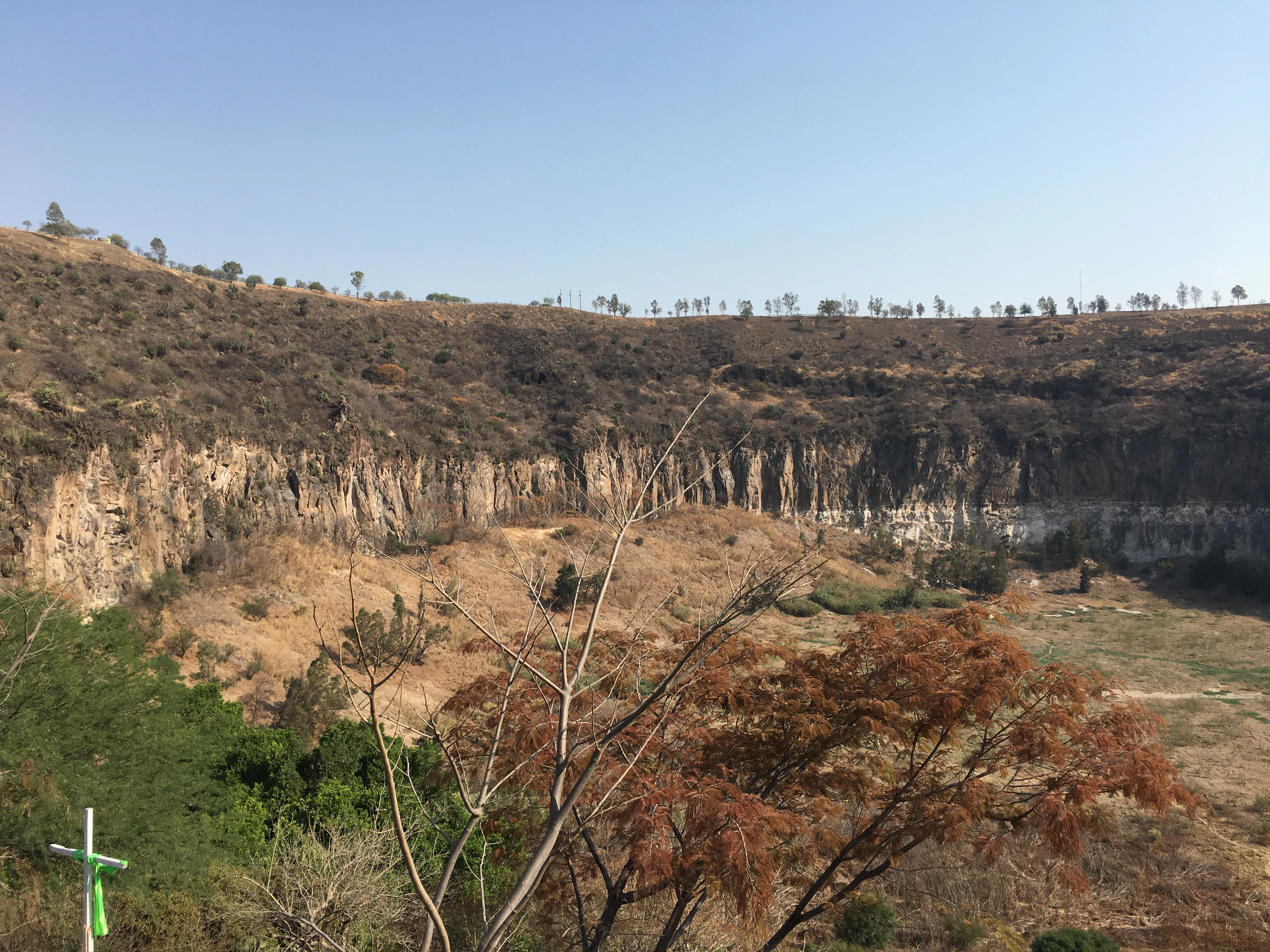 A wide view of a dry volcanic crater with steep rocky cliffs and sparse vegetation under a clear sky.
