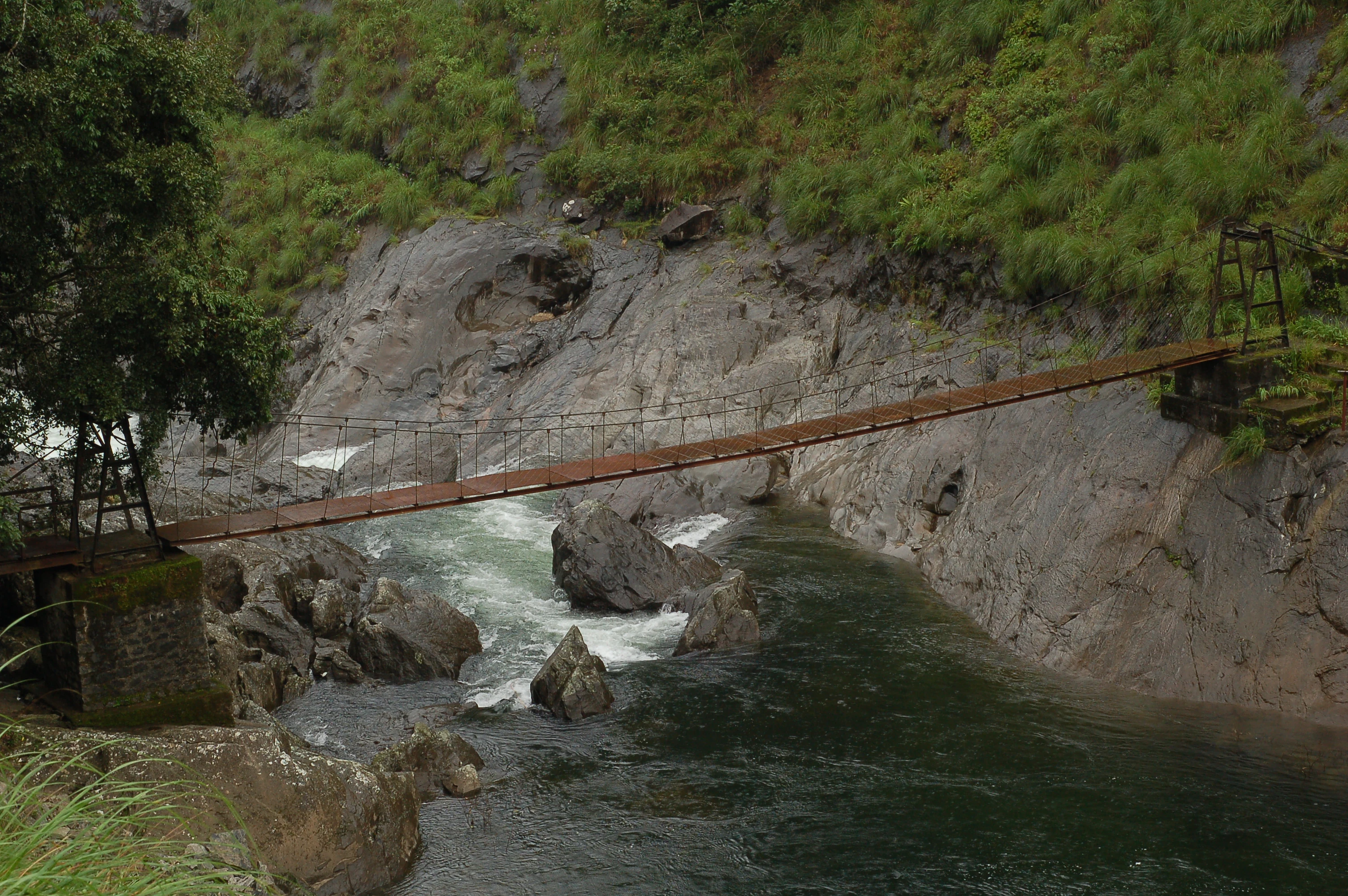 A suspension bridge spanning a river with rocky banks and dense green forest in the background