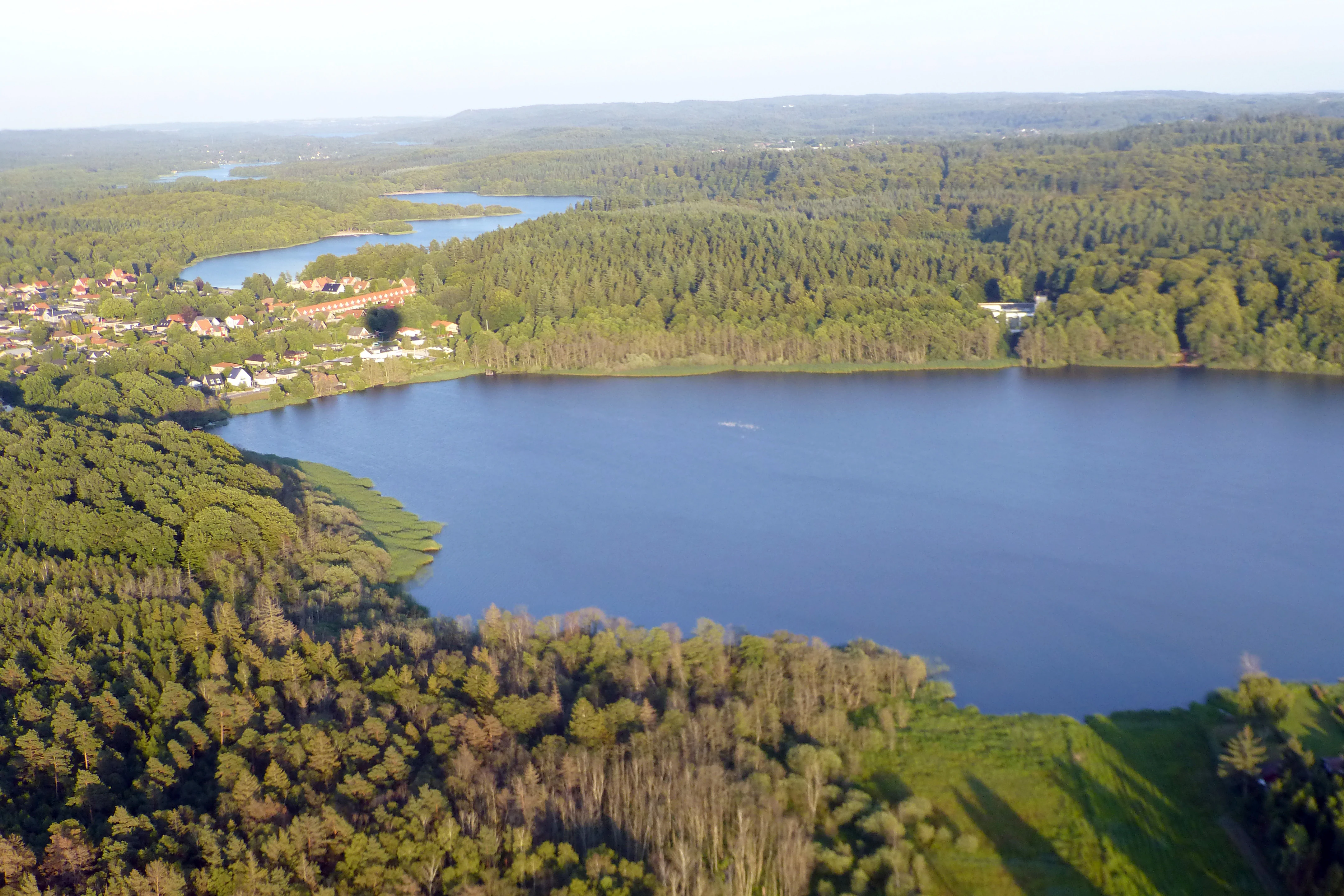 Aerial perspective looking down at a large lake surrounded by expansive green forests and a small residential area in Denmark.