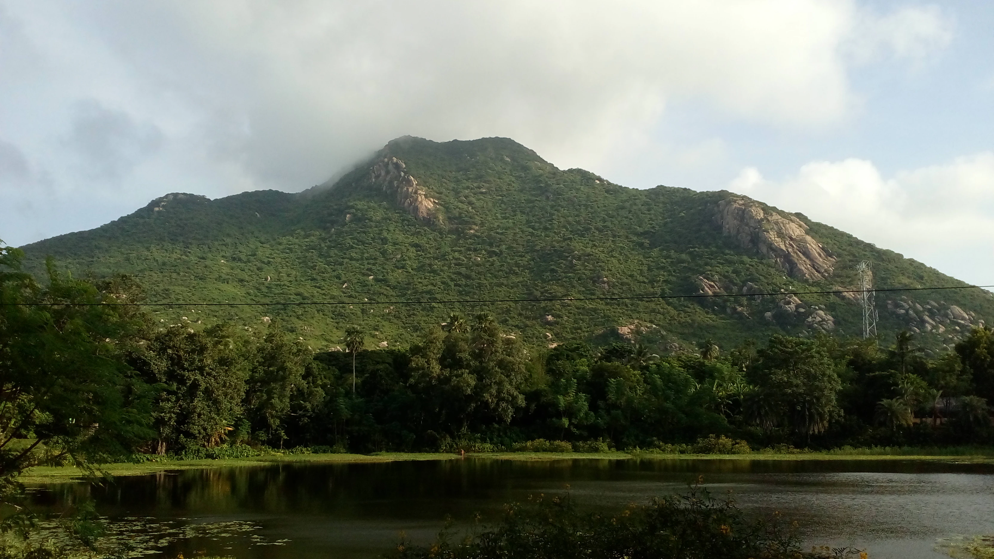 A mountain landscape with dense green vegetation, a calm lake in the foreground, and trees under a partly cloudy sky