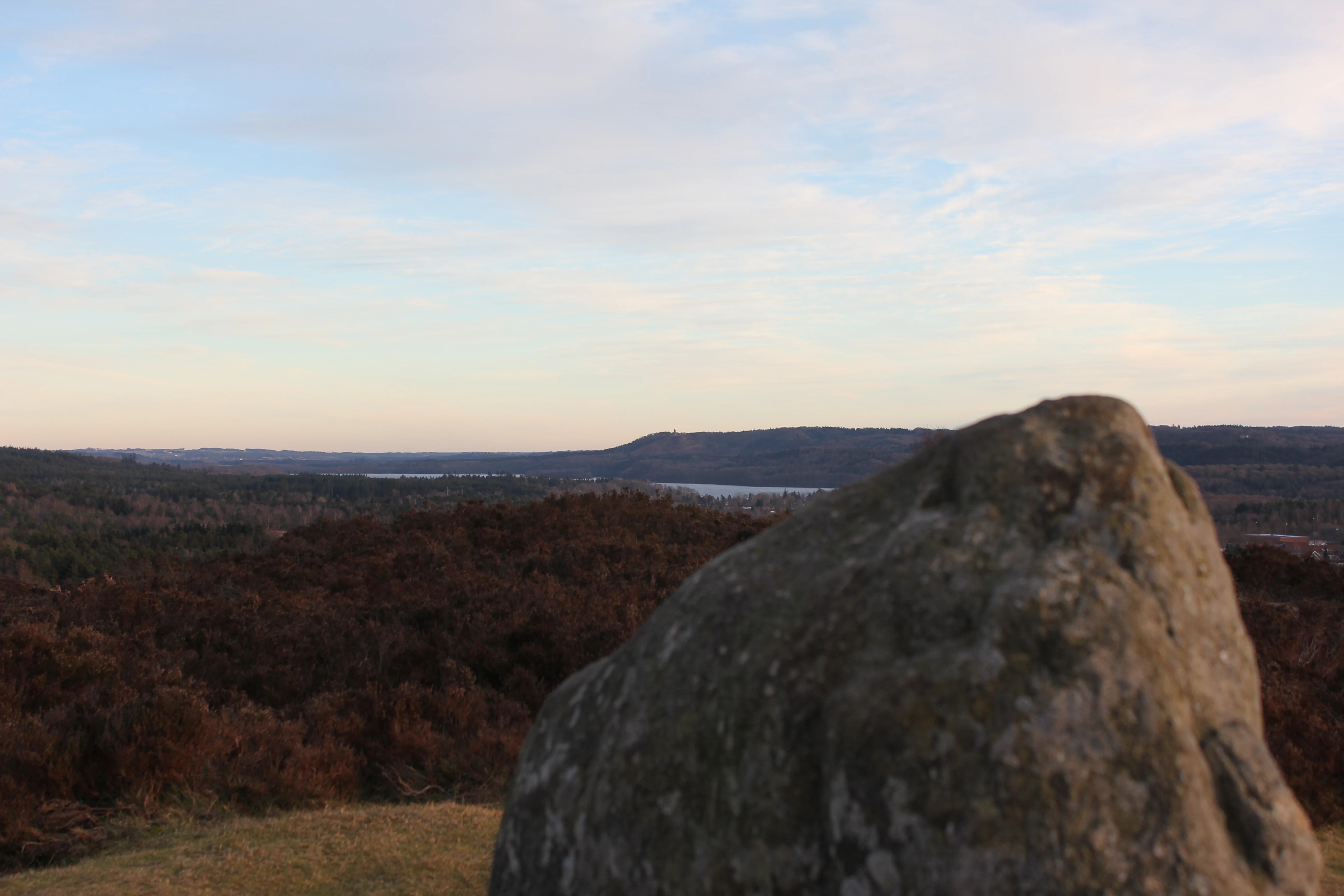 A large, weathered boulder sits in the foreground on a grassy hill, overlooking a rolling, dark-colored valley and a body of water under a pale evening sky.