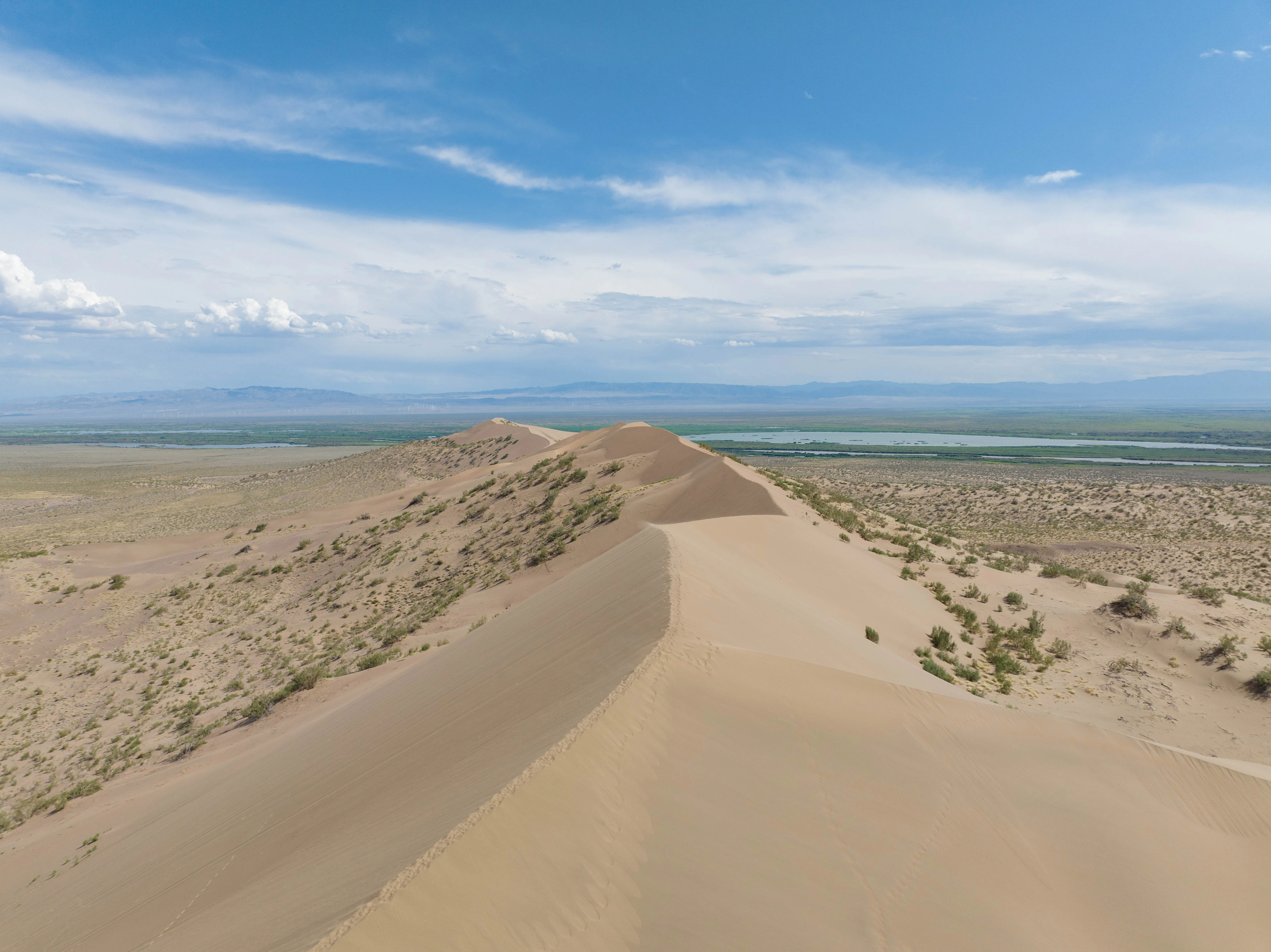 Una vista aérea que recorre la afilada cresta de una gran duna de arena, con vegetación árida y un horizonte lejano bajo un cielo azul nublado.