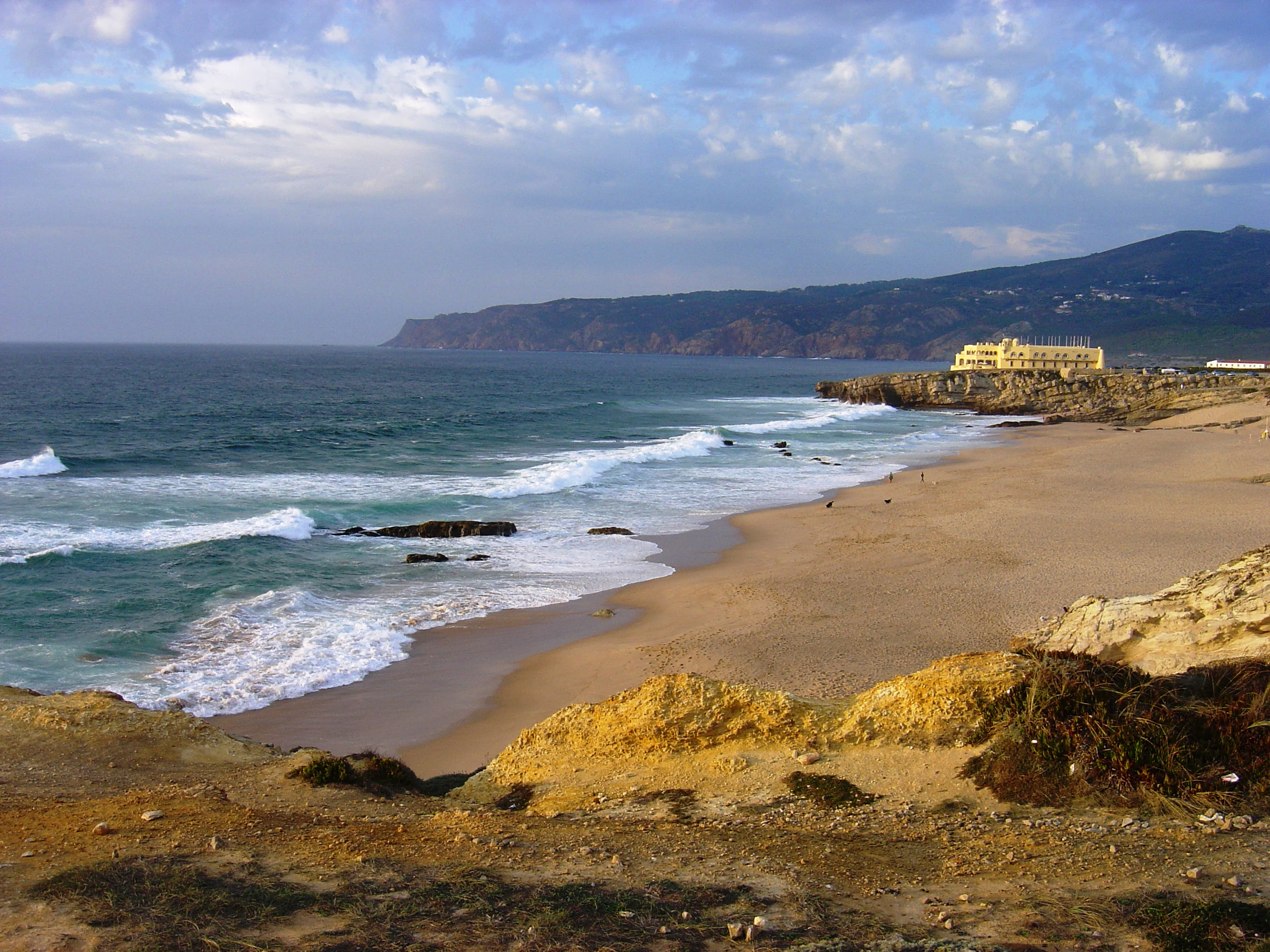 Sandy beach with turquoise waves crashing against rocky cliffs under a partly cloudy sky, distant buildings visible on the horizon