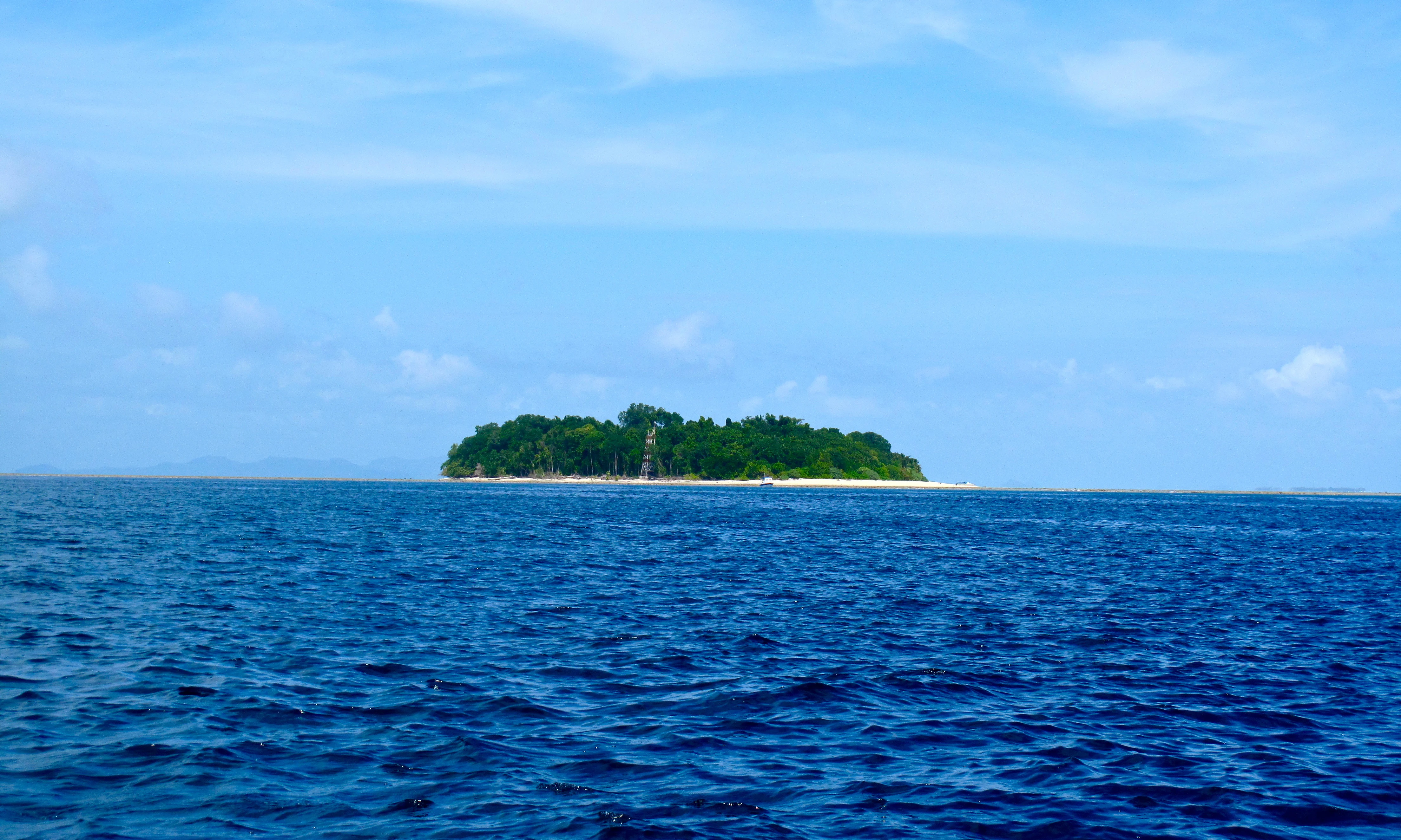 A small, tree-covered tropical island sits in the middle of a calm, deep blue sea under a light blue sky with faint clouds.