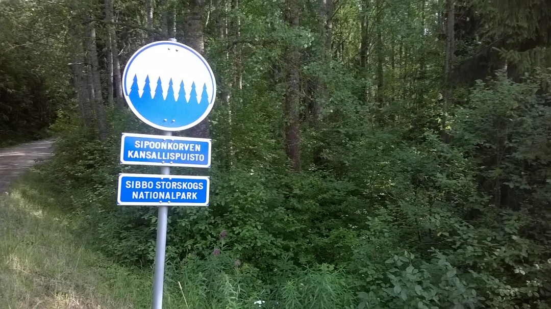 Blue and white sign on a pole with text 'Sipoonkorven Kansallispuisto' and 'Sipoonkorpi National Park' in a forest setting with green trees and a dirt path