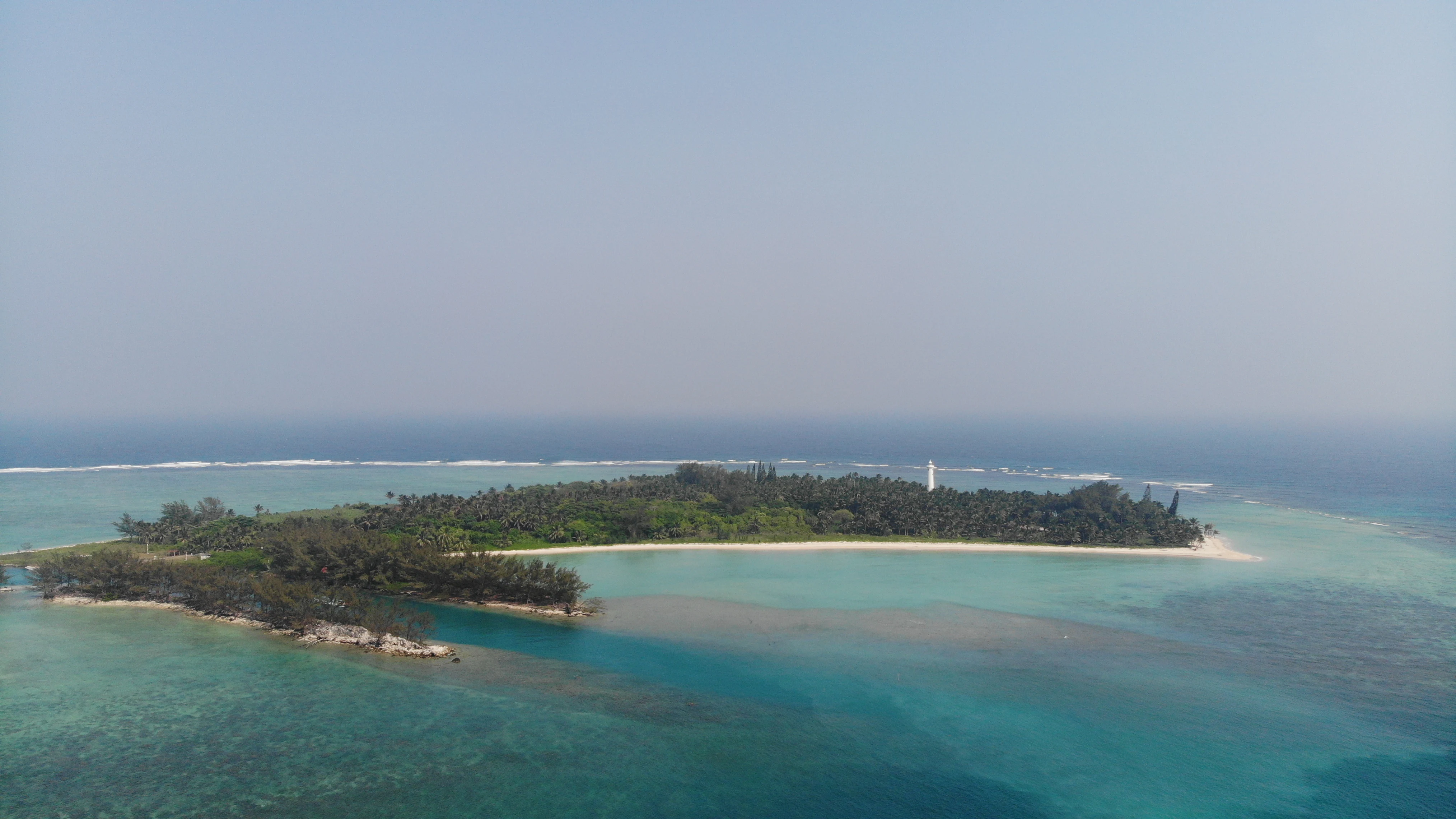 Una toma aérea de una pequeña isla boscosa rodeada de aguas poco profundas de arrecife turquesa y el océano azul profundo, con un faro blanco en la orilla.