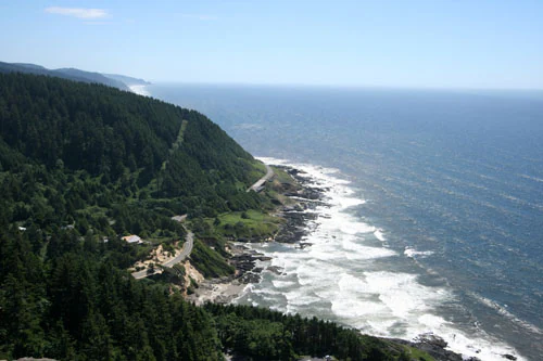 An elevated coastal view showing a dense evergreen forest meeting a rocky shoreline with crashing waves and a winding road following the coastline.