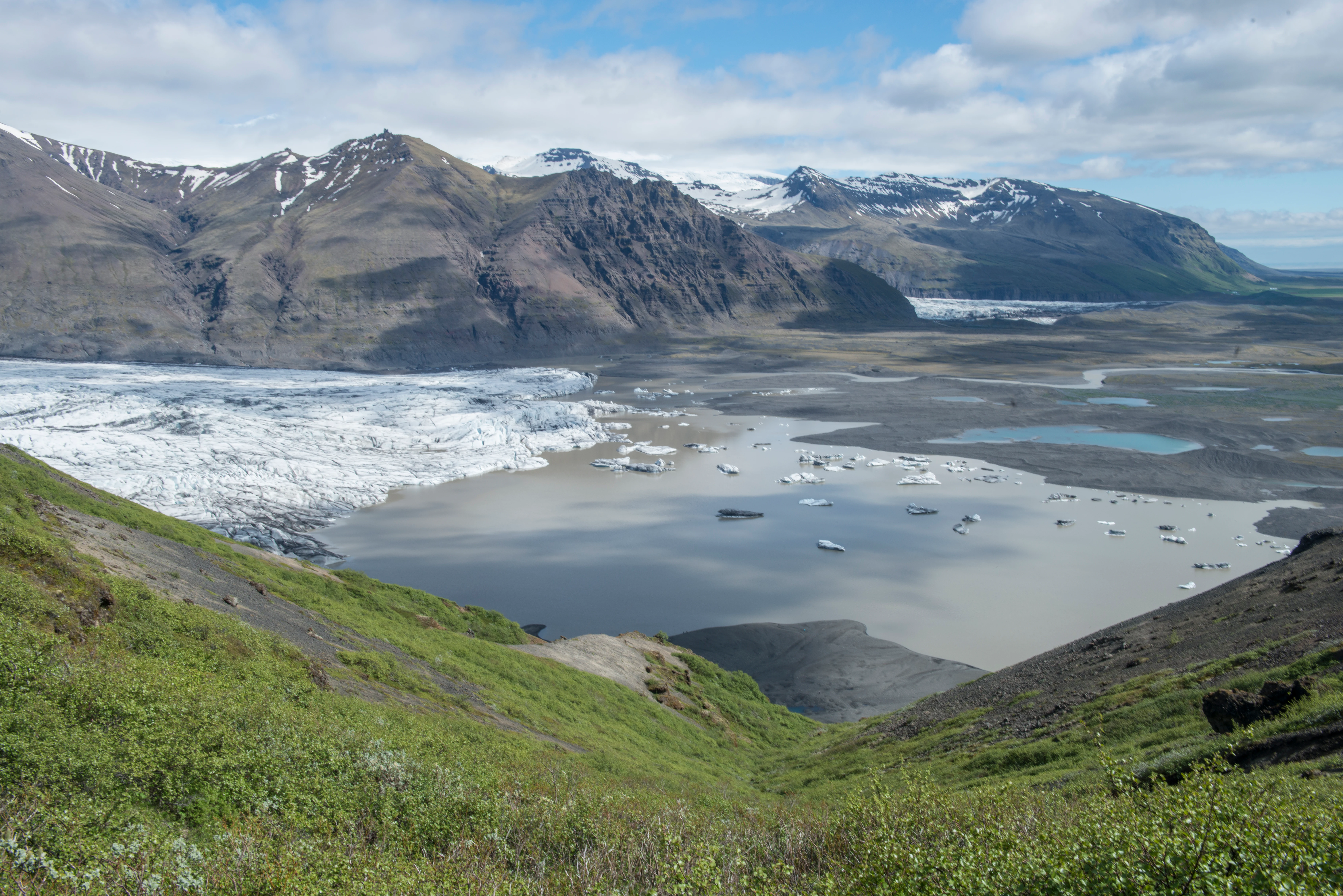 Panoramic landscape view showing glaciers, rivers, and mountain ranges with green vegetation in the foreground under a partly cloudy sky
